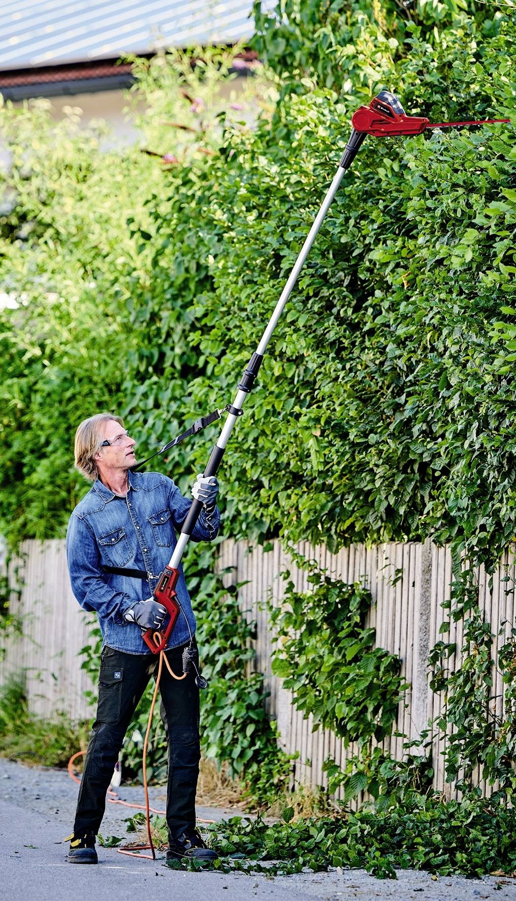 Egy személy elektromos sövényvágóval vág egy magas sövényt. Farmer dzsekiben és munkakesztyűben van.