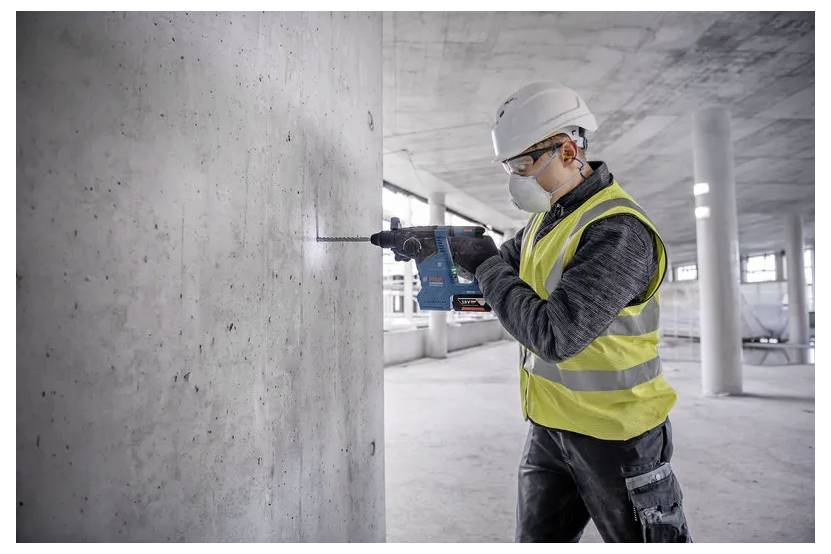 Egy munkavédelmi sisakot és védőmellényt viselő építőmunkás fúrógépet használ egy befejezetlen épület belső terében egy betonfalon.