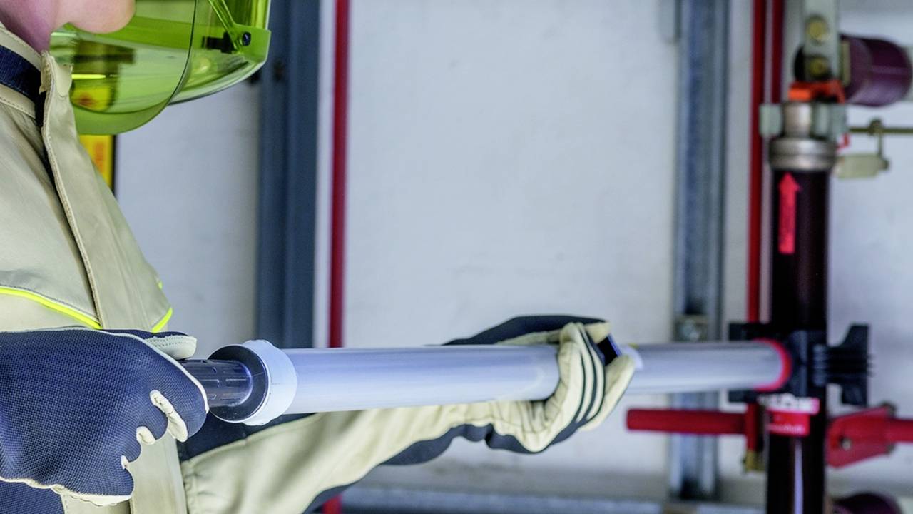 Védőruhát viselő személy csövet tart és egy vízvezetékrendszeren dolgozik egy technikai helyiségben.