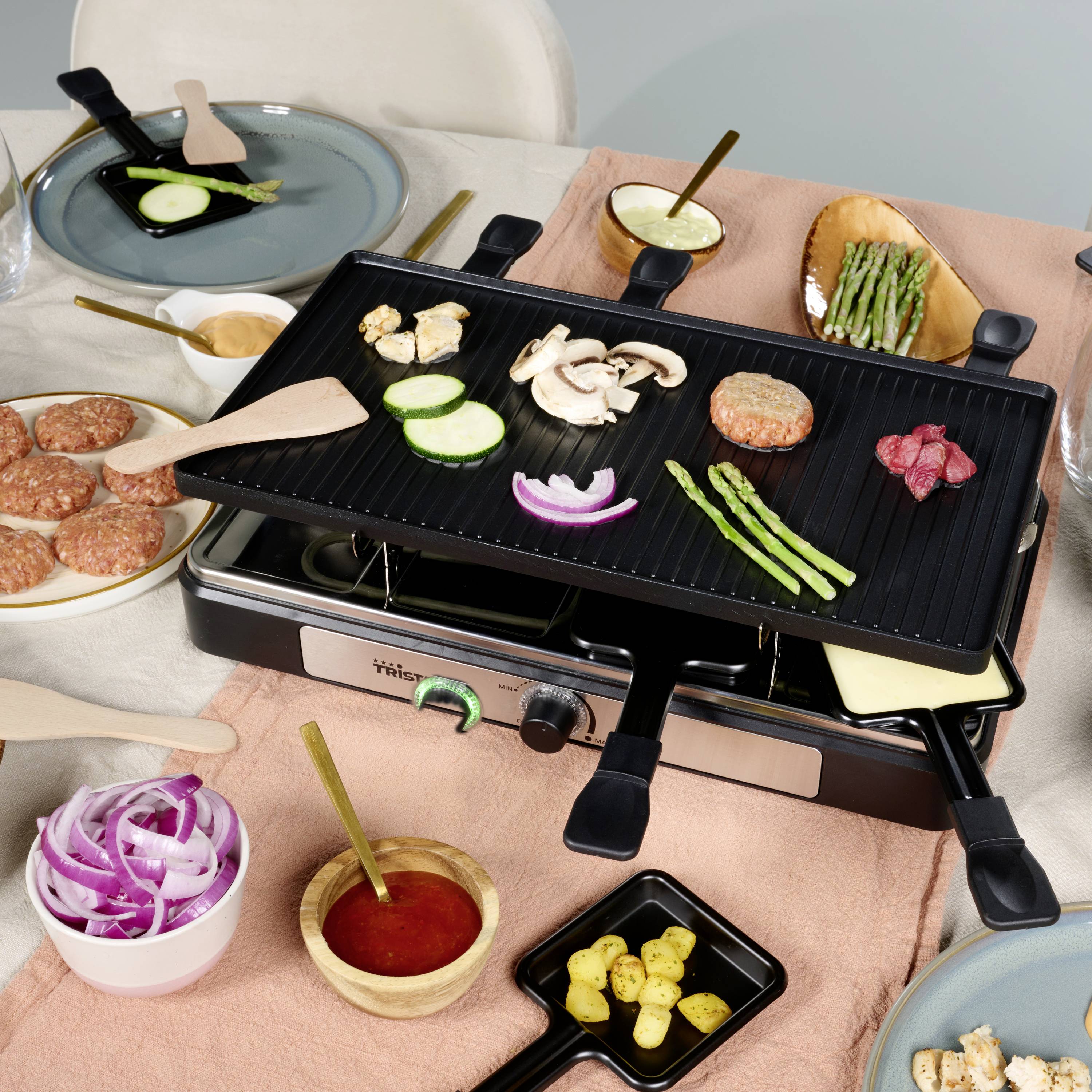 Elektromos asztali grill zöldségekkel és hússzeletekkel. Az asztalon emellett hamburgerpogácsák, hagymagyűrűk, szósz és krumplik fekszenek.
