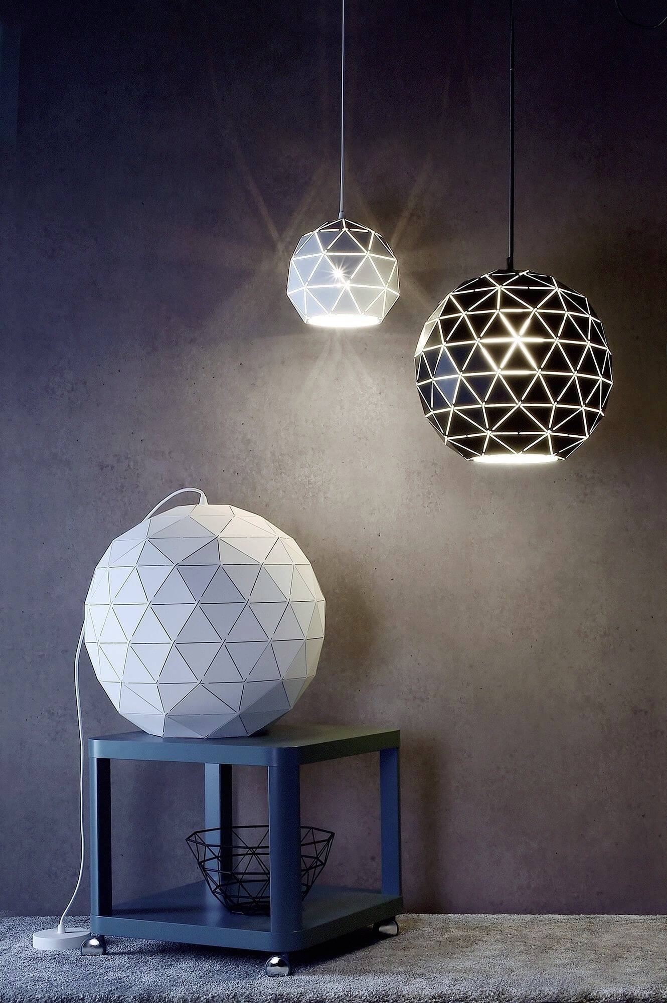Három modern geometriai mennyezeti lámpa lóg fehér és fekete színben egy szürke fal előtt. Egy lámpa az alatta lévő asztalon áll.