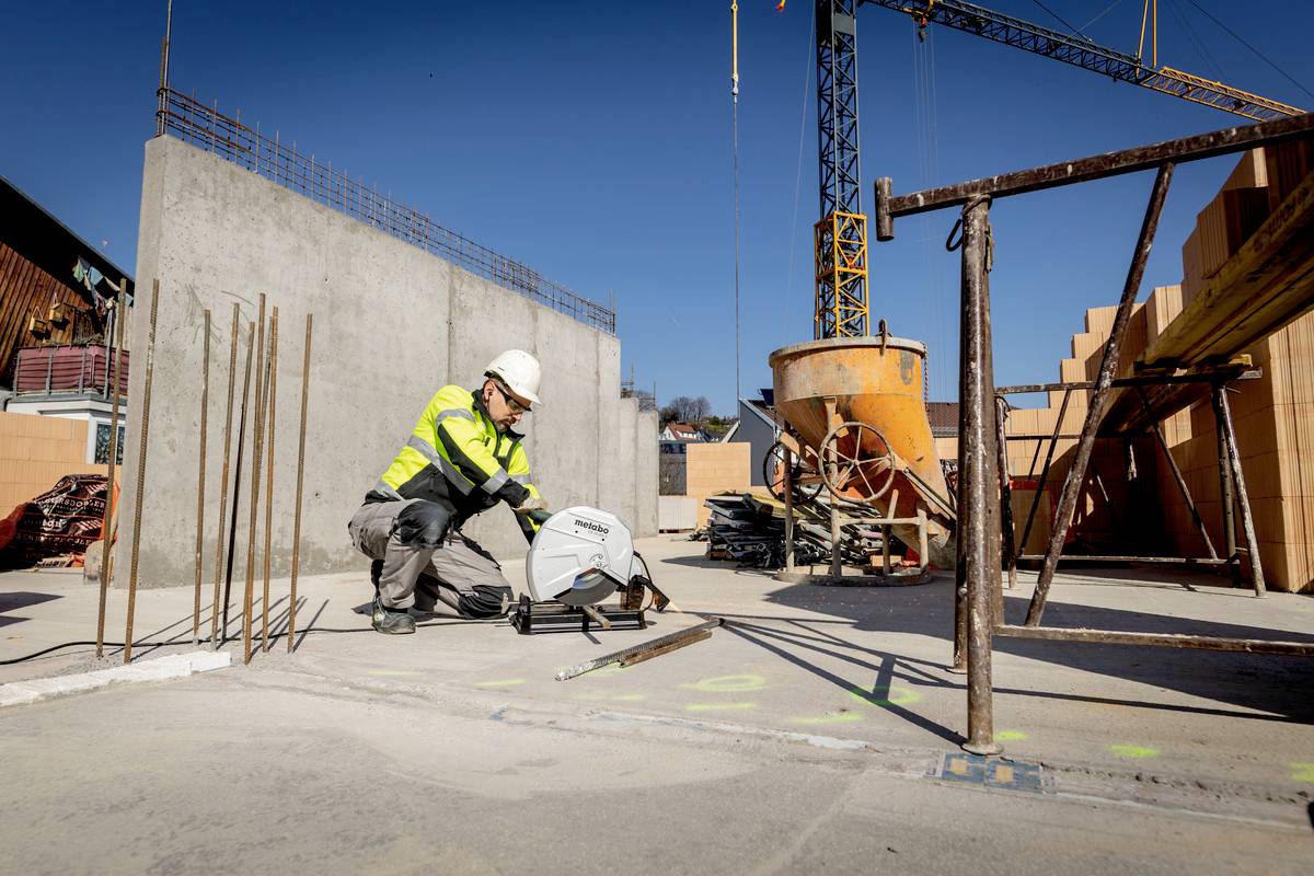 Egy védőfelszerelést viselő munkás vágógépet használ egy építkezésen. A háttérben betonszerkezetek és egy daru látható.
