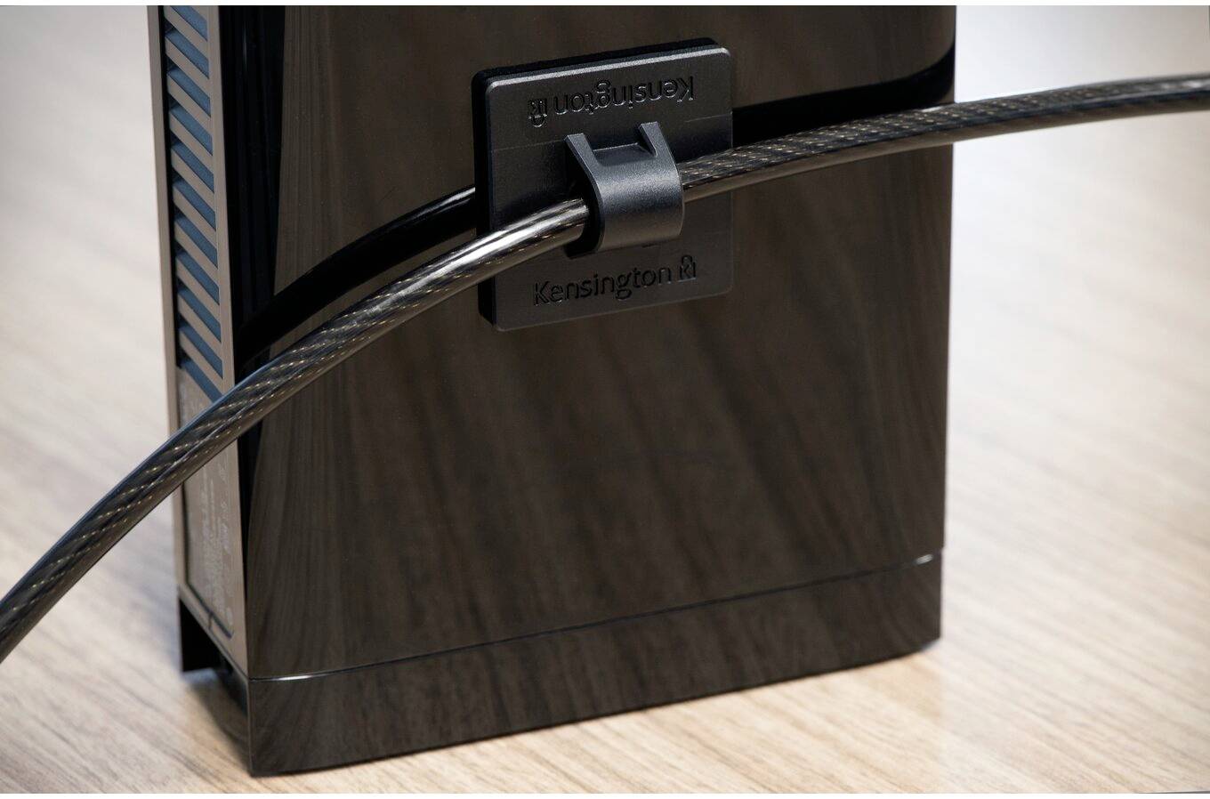 Egy fényes, fekete eszköz áll egy asztalon. Egy biztonsági kábel egy Kensington-zárral van csatlakoztatva, és rögzíti a készüléket.