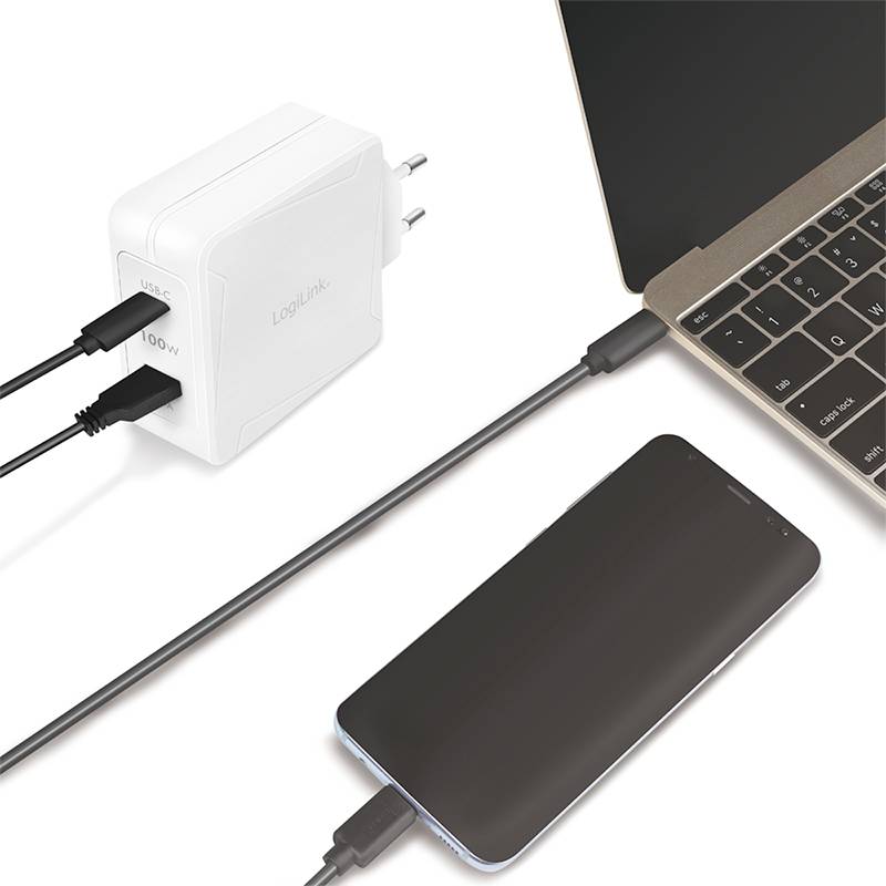 Egy okostelefon fekete kábellel egy fehér fali töltőadapterhez csatlakozik. Mellette egy nyitott laptop fekszik.