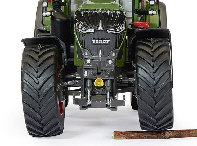 Egy zöld traktor nagy kerekekkel halad a szabadban. A traktor előtt egy kis faágdarab fekszik.