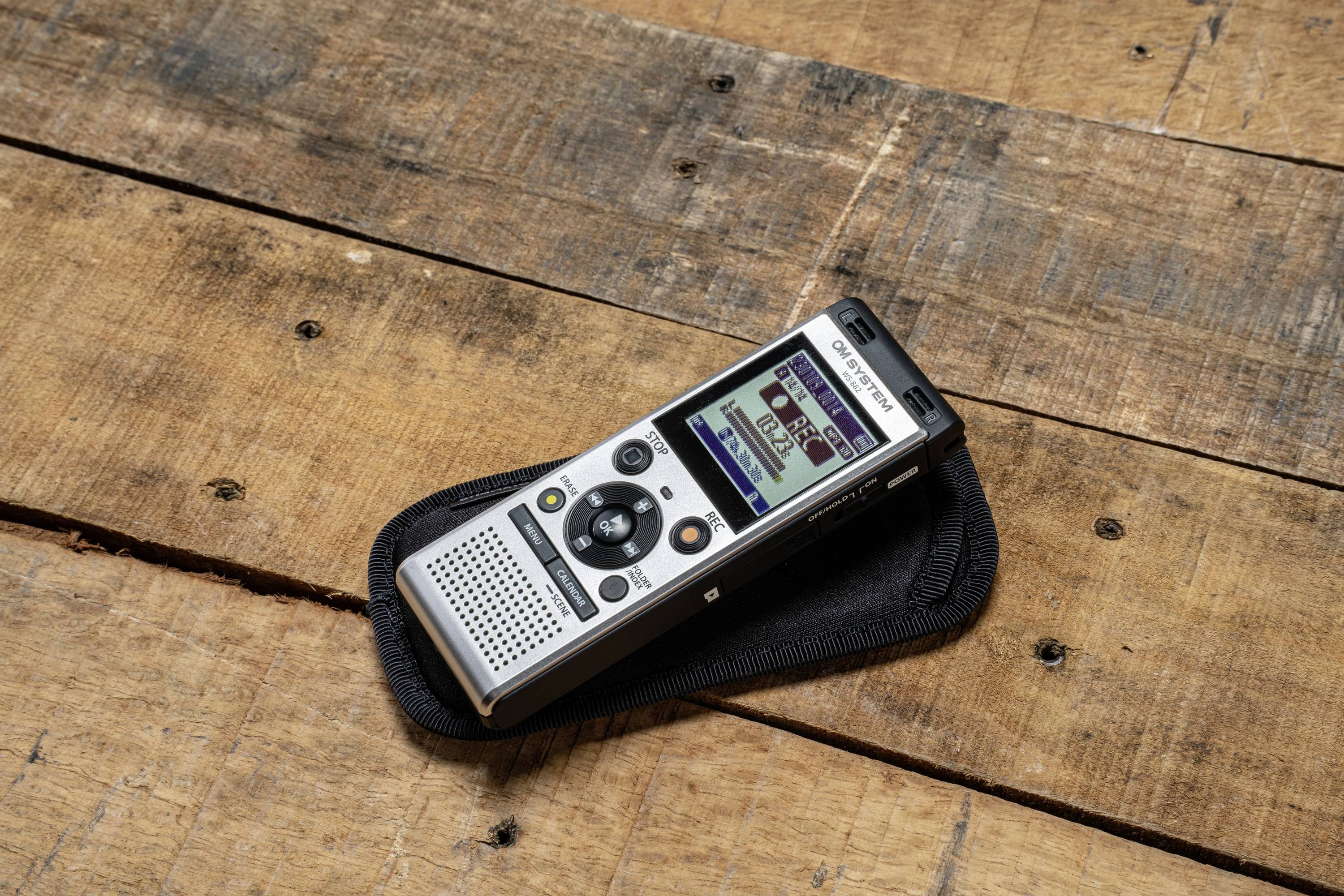 Hordozható diktafon kijelzővel, ami egy fa asztalon fekszik. A készülék felvételi funkciót mutat, hangfelvételek rögzítésére használják.