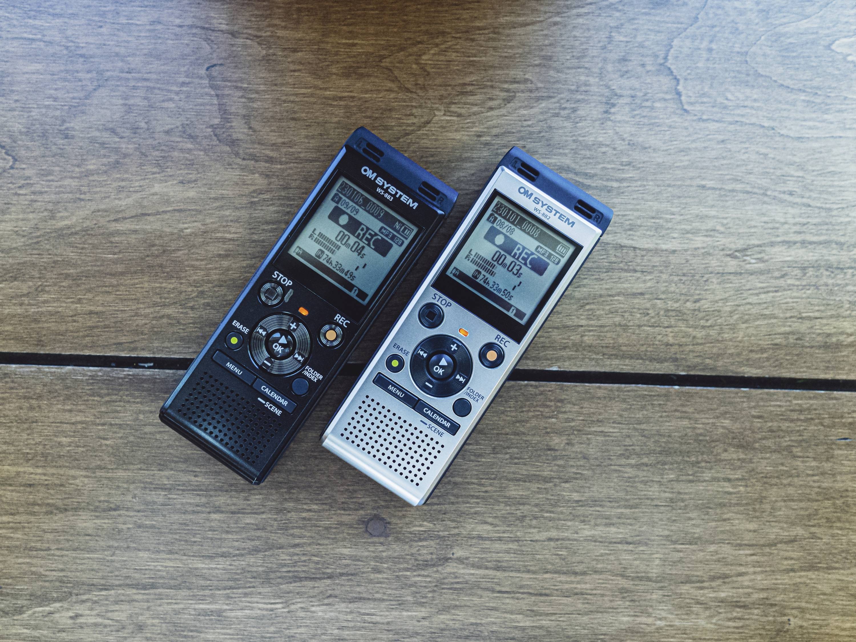 Két diktafon fekszik egymás mellett egy fa asztalon. Az egyik fekete, a másik ezüst. Mindkettőn LCD-kijelző látható a felvételi idővel.