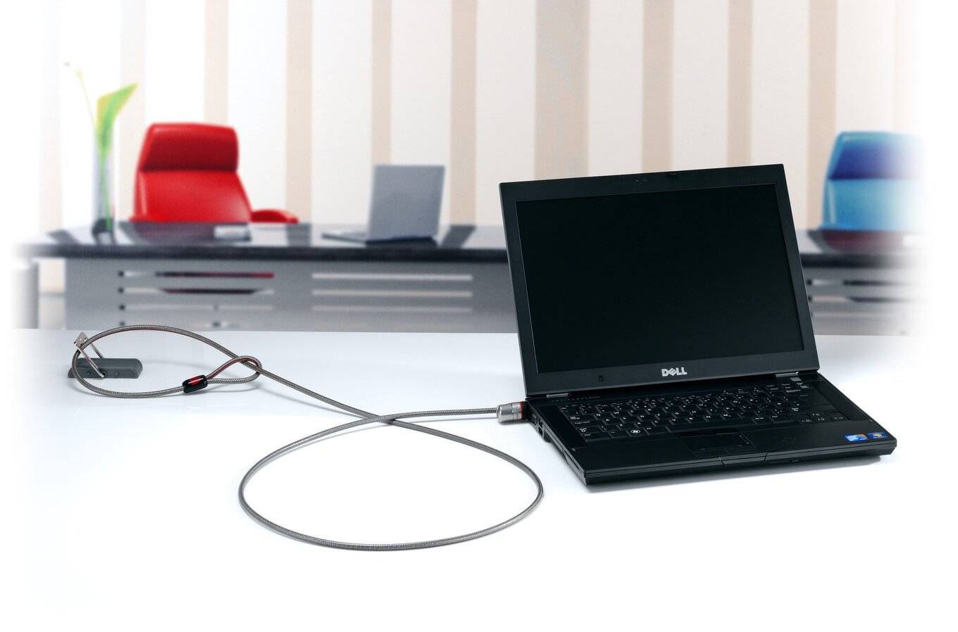 Egy laptop biztonsági kábellel van rögzítve egy íróasztalhoz. A háttérben székek és további laptopok láthatók.