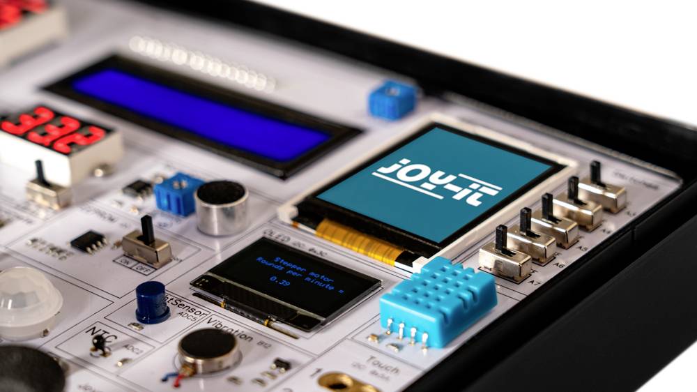 Elektronikus kísérleti panel különböző kapcsolókkal, gombokkal, LED-ekkel és egy kisméretű kijelzővel, rajta a 'joy-it' logóval.
