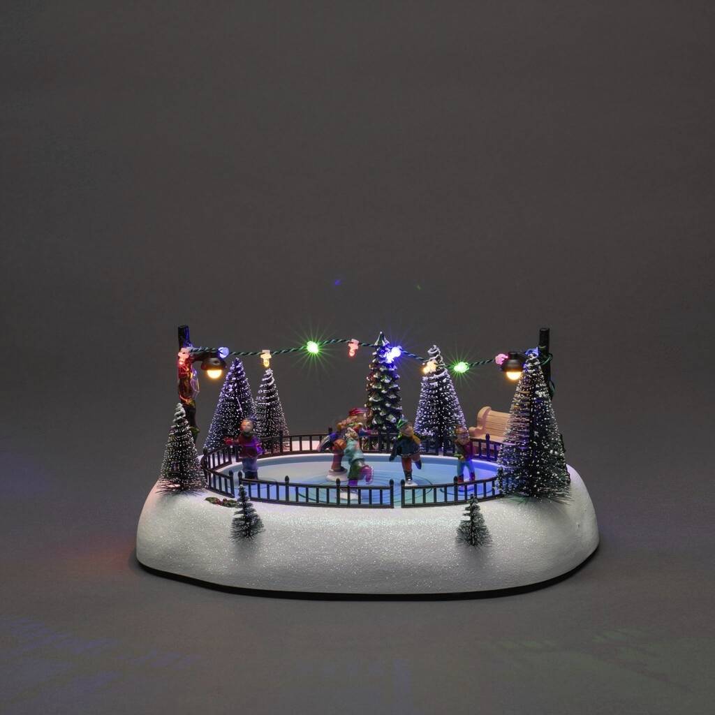 Egy miniatűr karácsonyi falu korcsolyapályával. Figurák korcsolyáznak a kivilágított fenyőfák között. Dekoratív fényfüzérek a szegélyén.