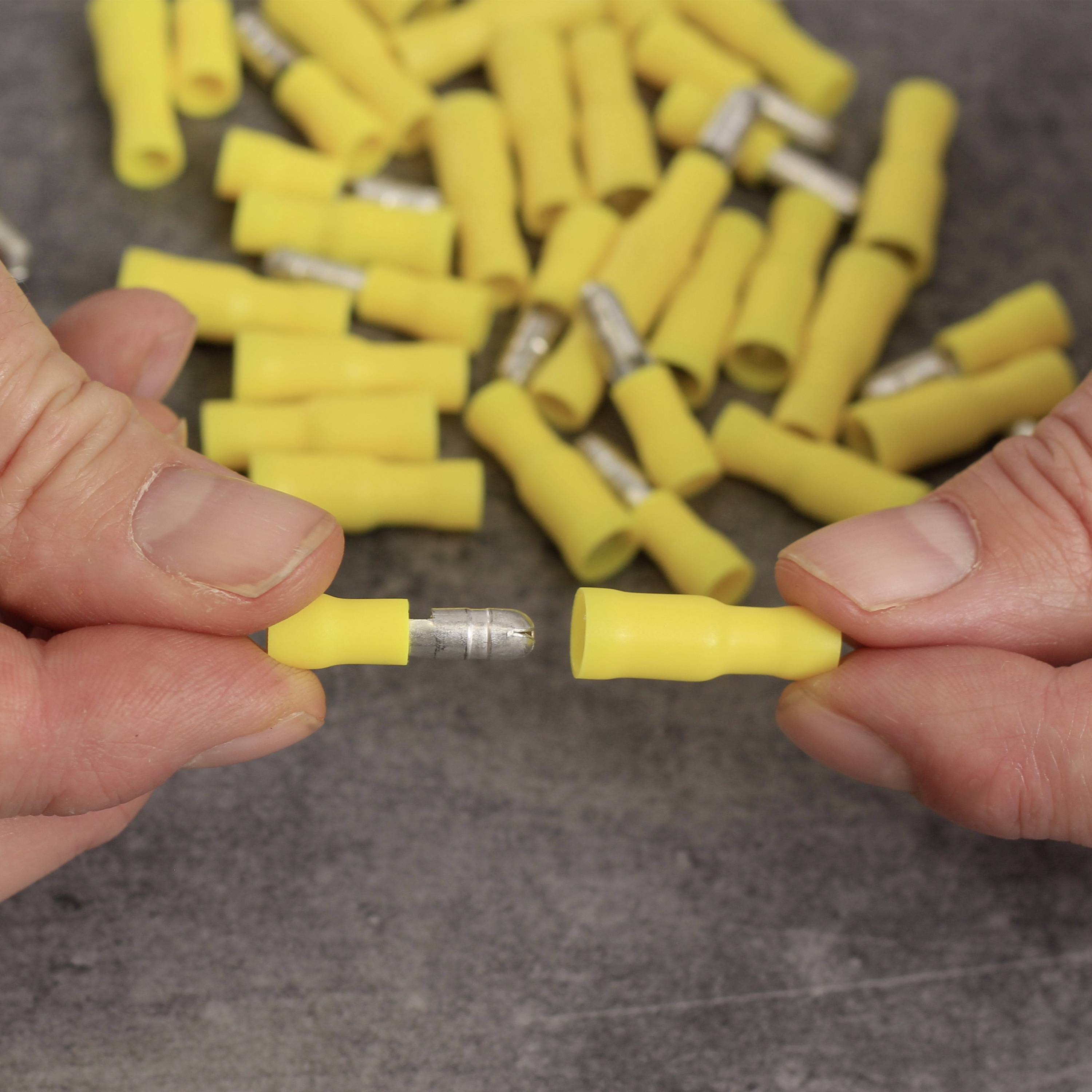 Két kéz sárga elektromos kábel csatlakozókat tart egy szürke felületen; több csatlakozó látható a háttérben.