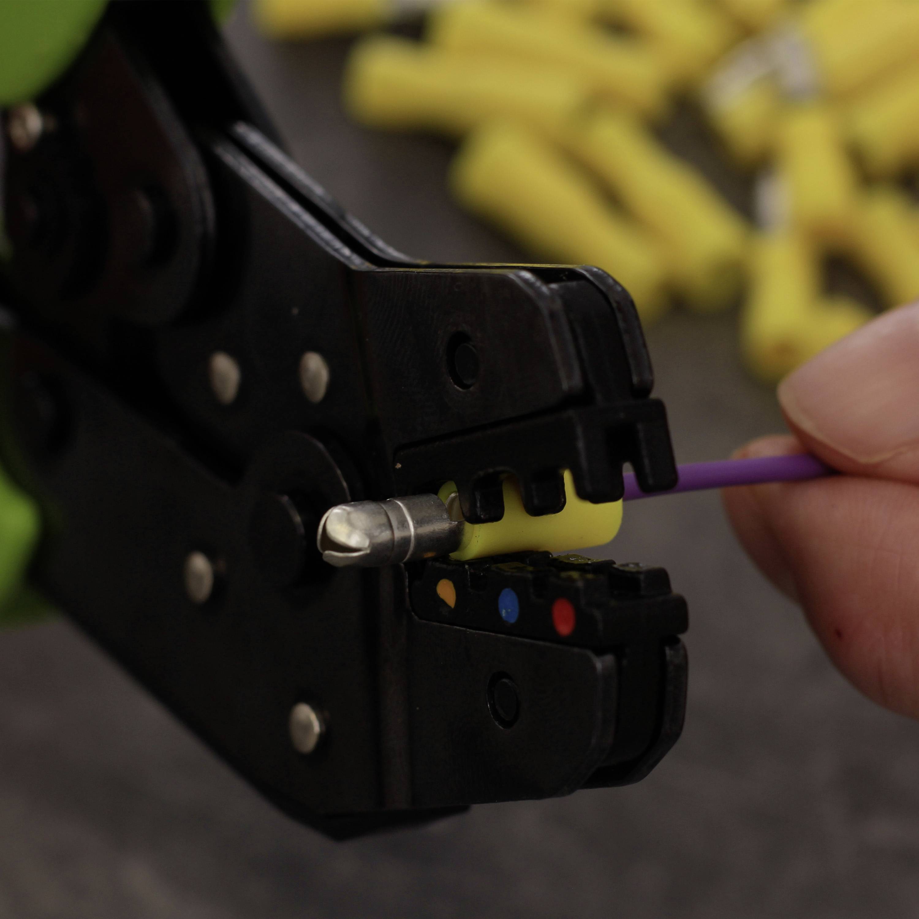 Egy kéz krimpelőfogót használ egy lila kábel sárga kábelsaru csatlakoztatásához. A háttérben elmosódott sárga alkatrészek láthatók.