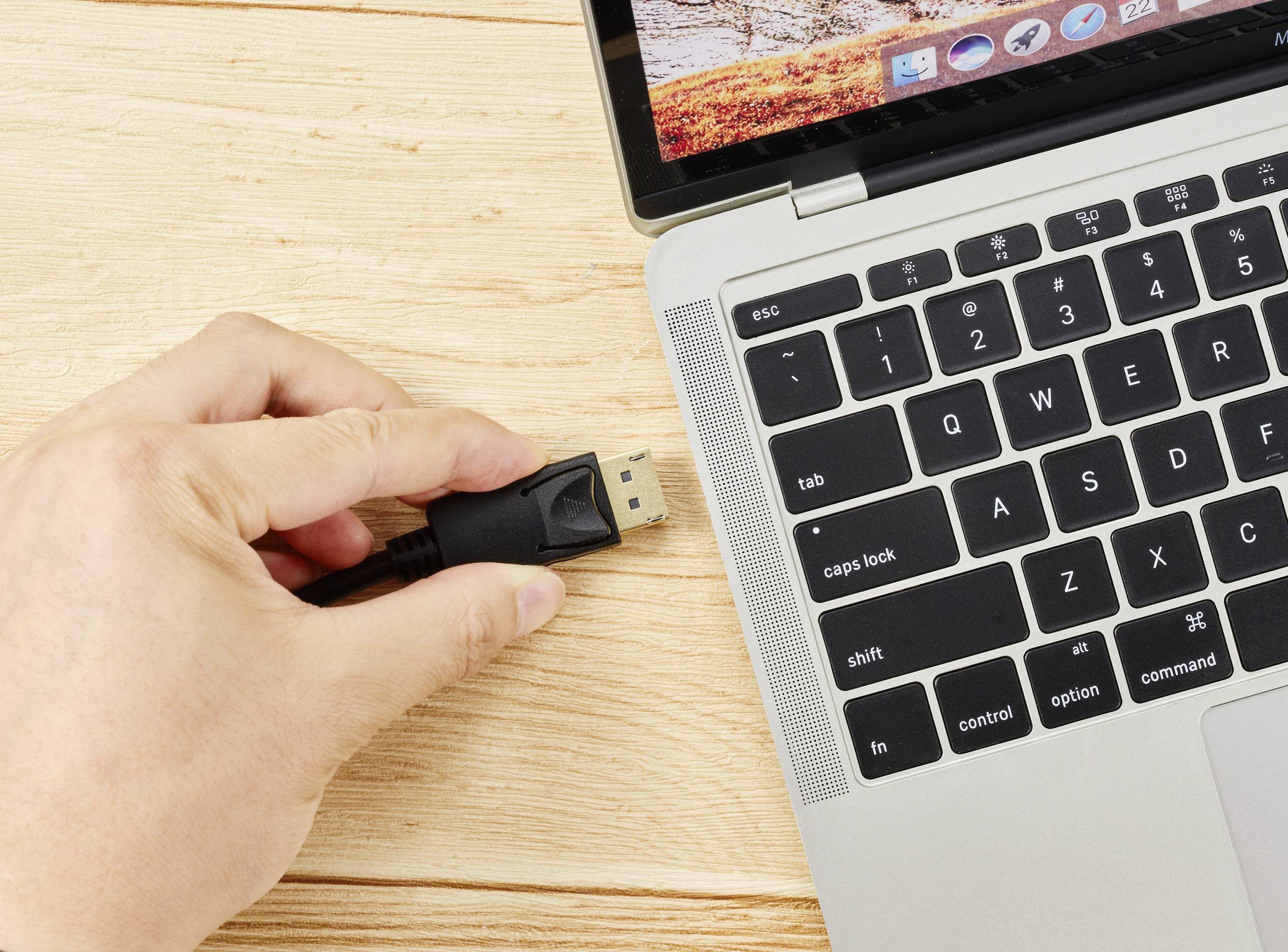 Egy kéz HDMI-kábelt tart egy laptop mellett. A laptop nyitott billentyűzettel és egy természetes háttérrel rendelkező képernyővel rendelkezik.