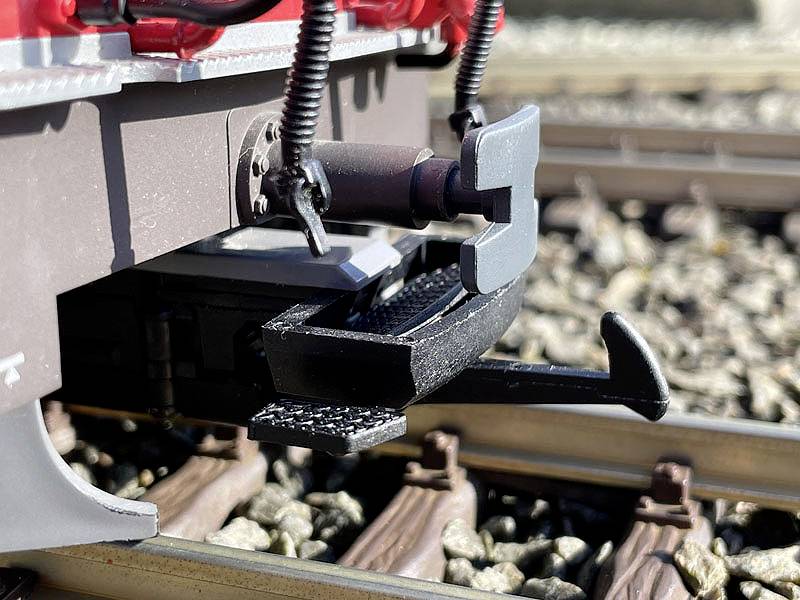 Egy modellvasút-szerelvény csatlójának közeli felvétele. A csatolószerkezet részletei és a kavicságy látható a háttérben.