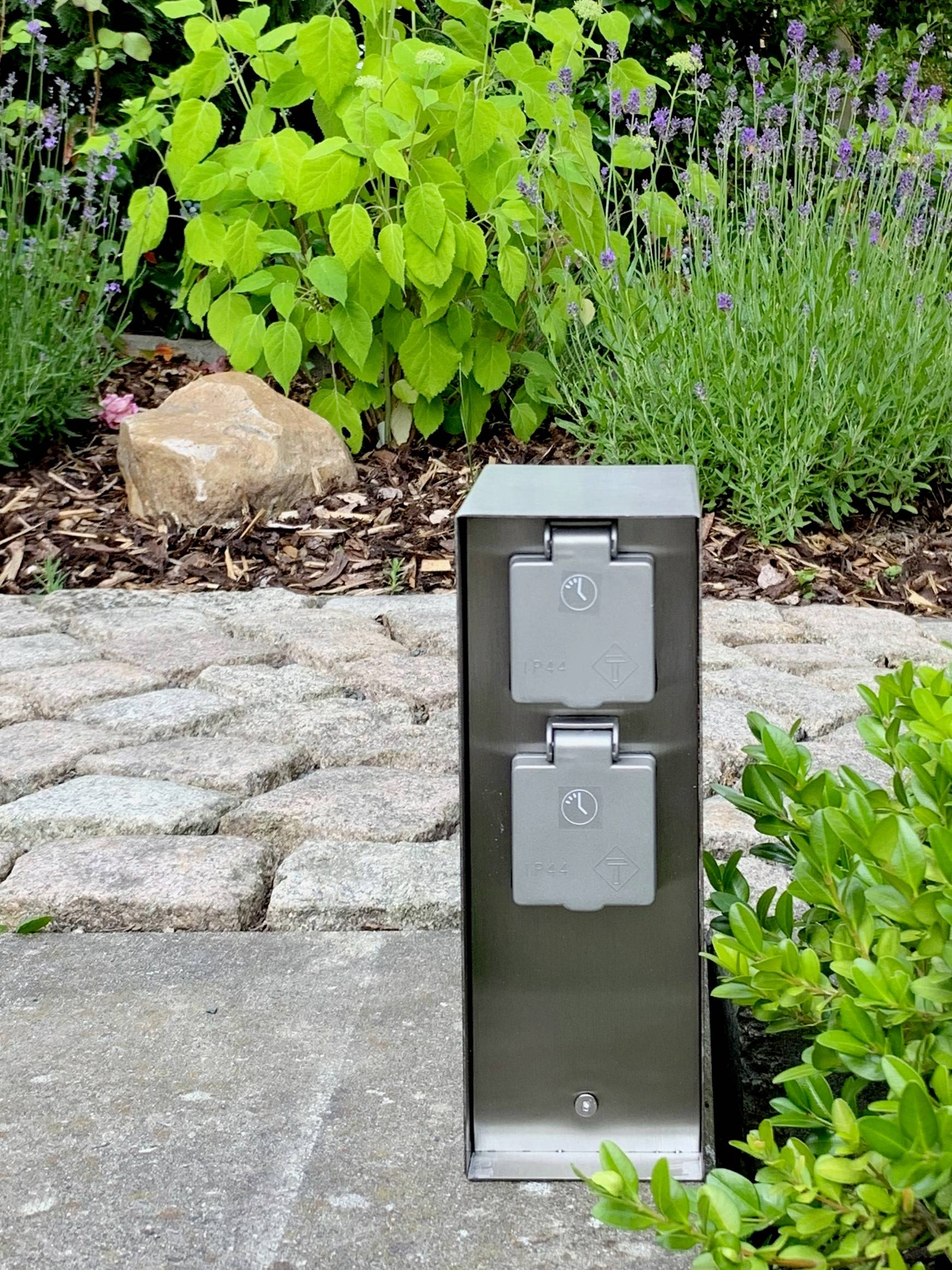 Fém elektromos csatlakozódoboz a kertben, térburkolatos út és növények között elhelyezve.