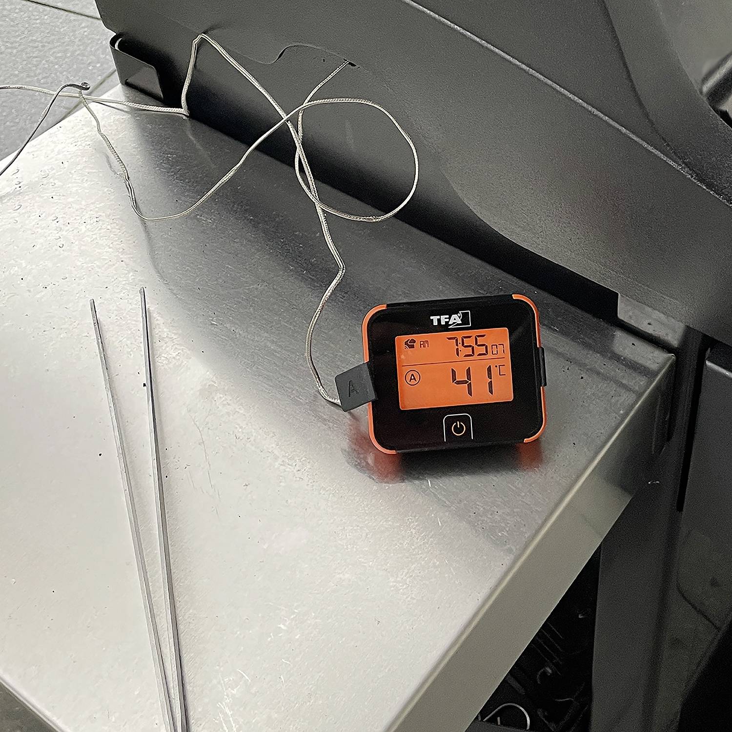 Egy 41°C-os hőmérséklettel leolvasható hőmérő egy fémfelületen fekszik. Két szonda van a készülékhez csatlakoztatva.