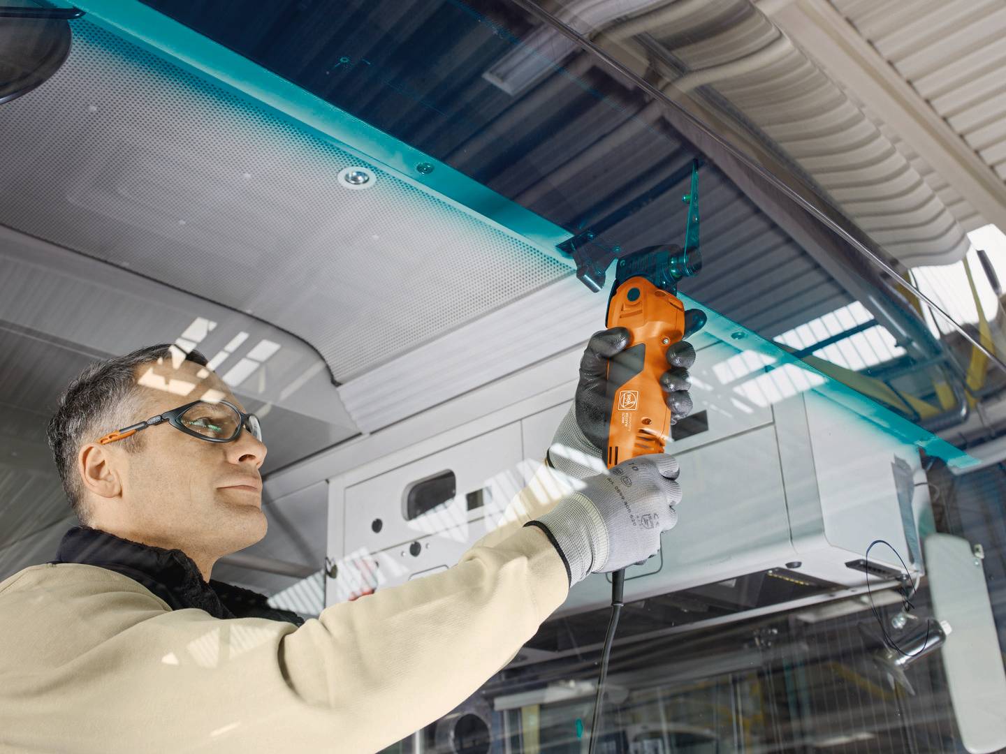 Egy védőszemüveget viselő személy egy narancssárga elektromos szerszámmal javít vagy karbantart egy járműtetőt egy műhelyben.