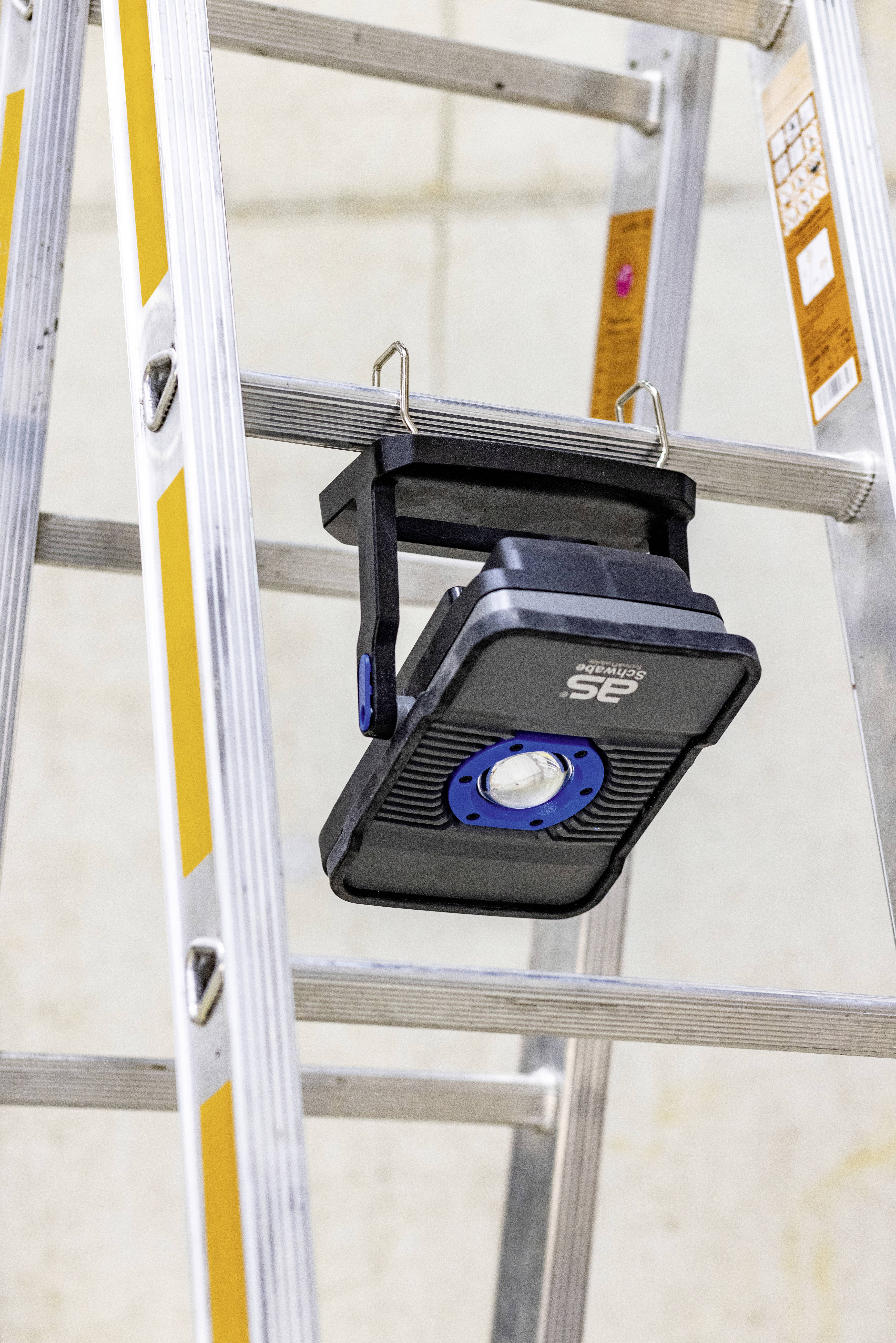 Egy hordozható LED-lámpa egy alumínium létrára van rögzítve. A lámpa lefelé néz, és kapcsok tartják.