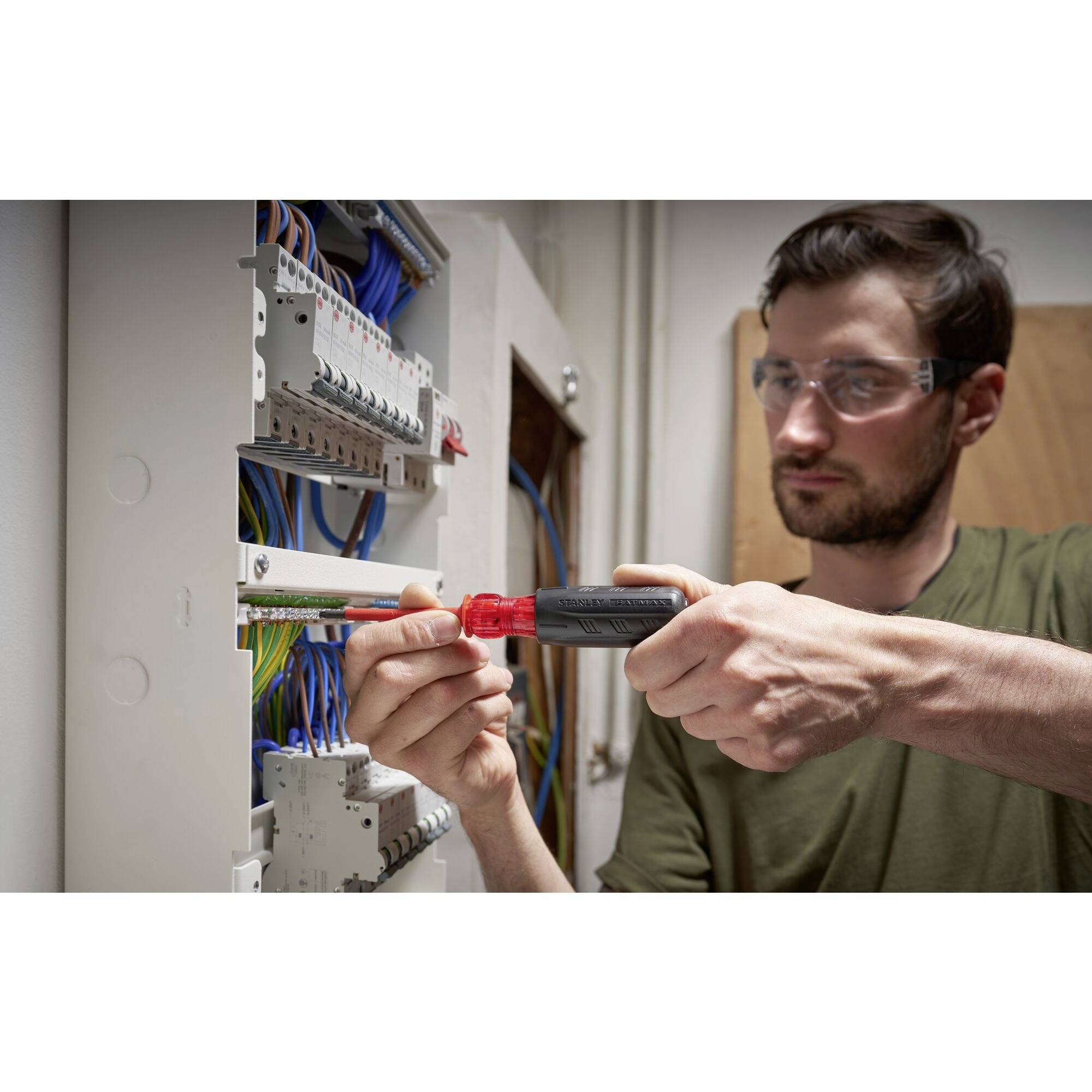 Egy védőszemüveget viselő férfi egy nyitott elektromos elosztószekrénynél ellenőrző készüléket használ, és vizsgálja az elektromos csatlakozásokat.