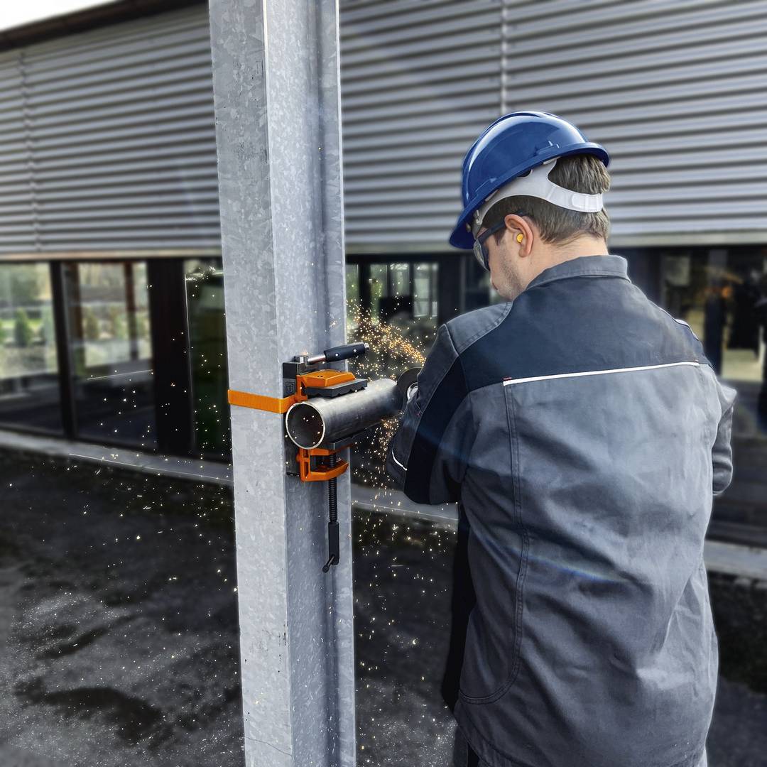 Egy védősisakot és fülvédőt viselő munkás egy szabadtéri állványon fémcsövet vág, miközben szikrák röpködnek.