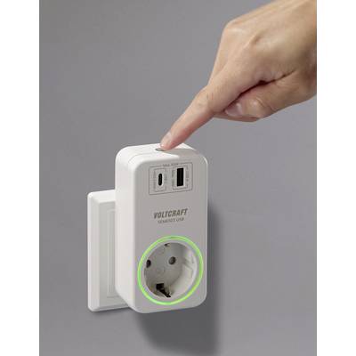 Egy kéz egy fehér elosztóadapterre mutat, amely USB-csatlakozókkal és zöld LED-gyűrűs jelzéssel rendelkezik, és a falon van elhelyezve.