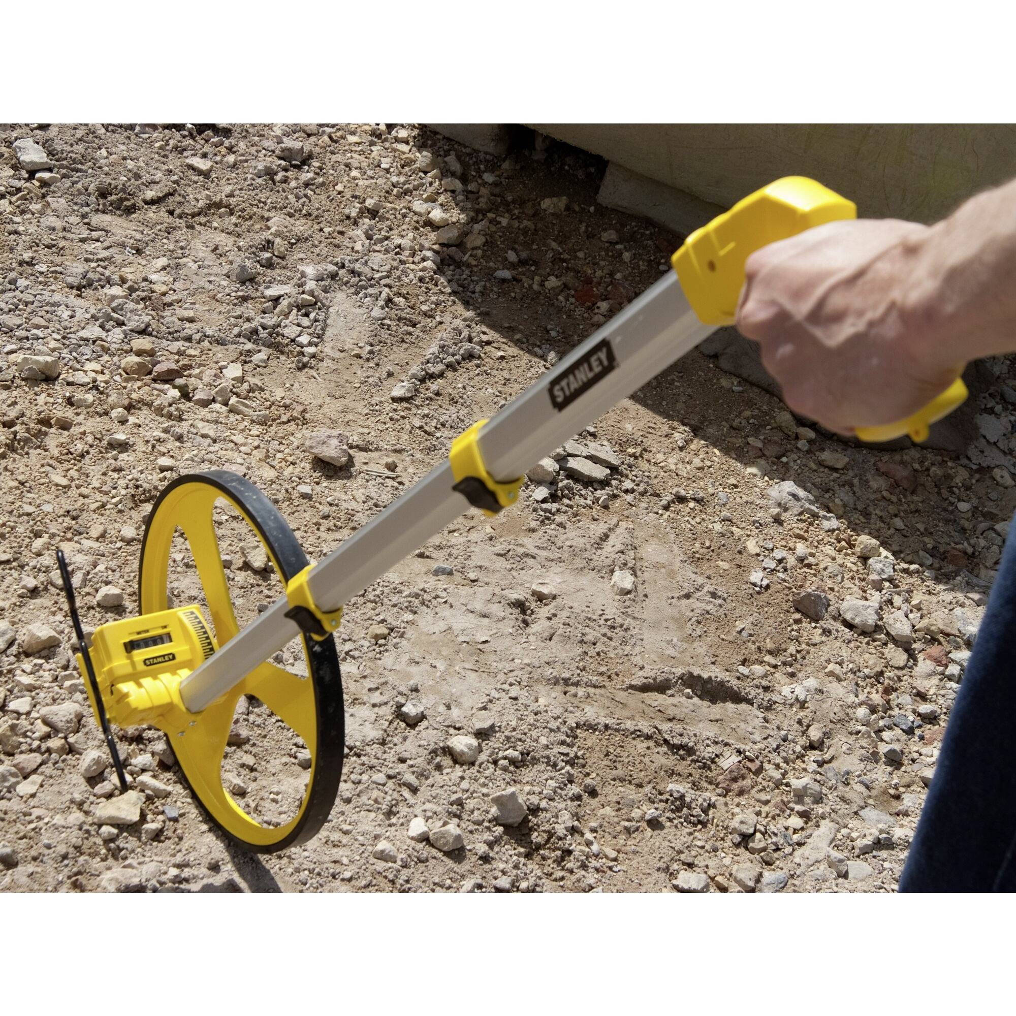 Egy kéz egy sárga mérőszalagot tart homokos talajon. Az eszközt távolságok mérésére használják.