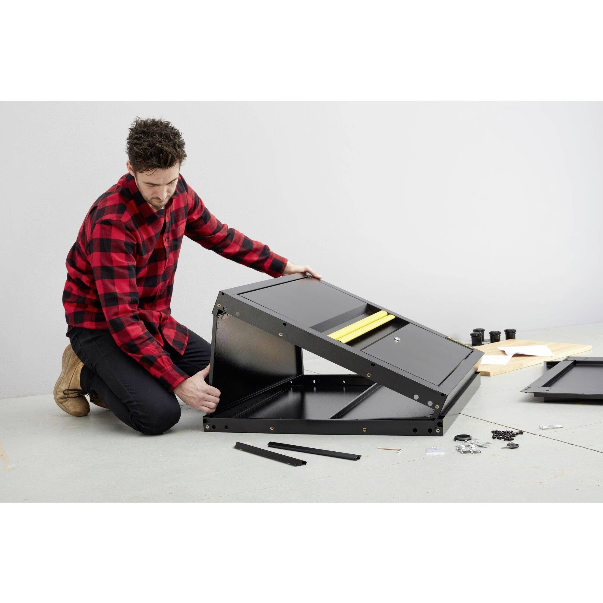Egy kockás ingű férfi egy fekete bútordarabot szerel össze. Világos padlón áll, körülötte szerszámok és alkatrészek hevernek.