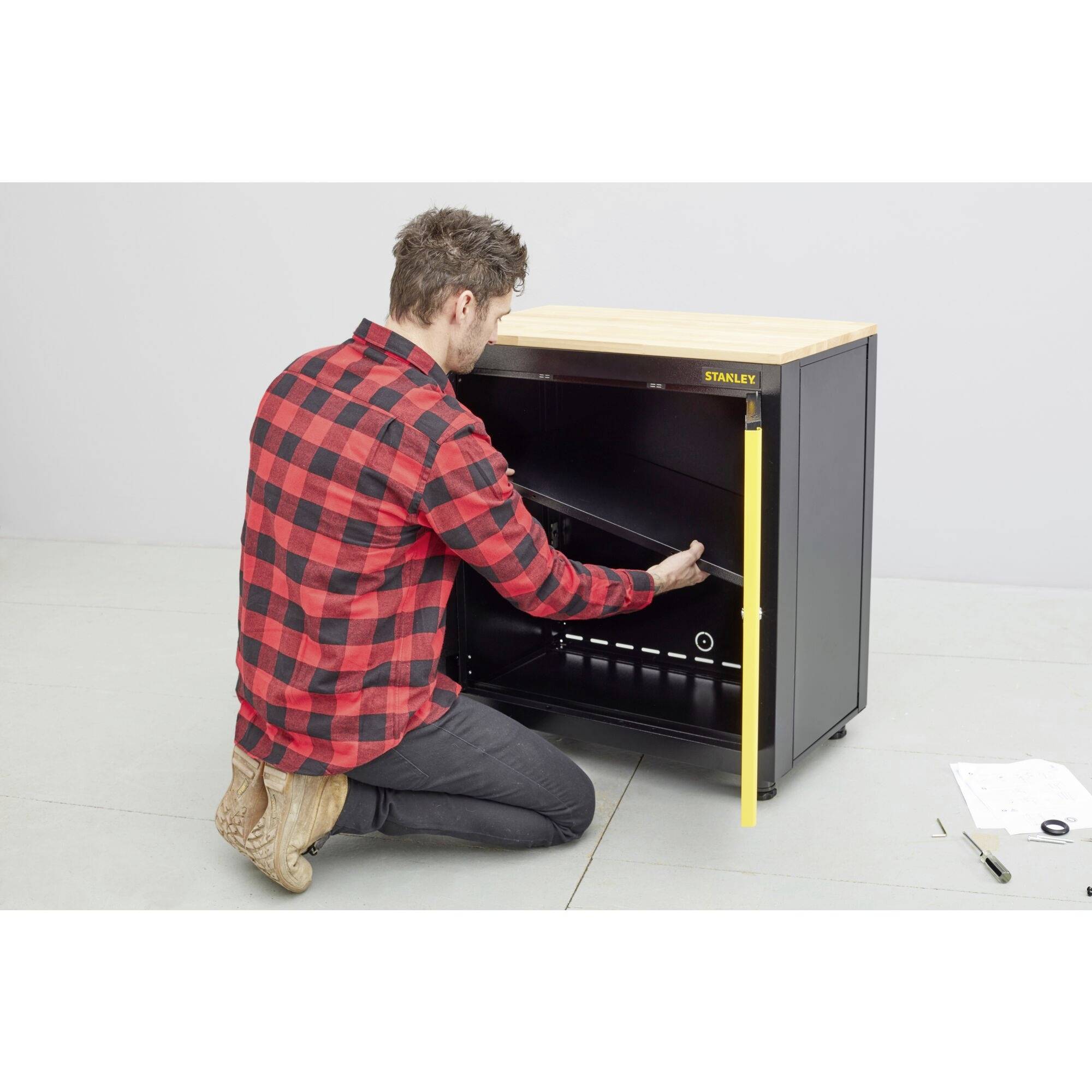 Egy férfi egy fekete házat szerel össze, térdel a padlón, kockás inget visel. Szerszámok és útmutató fekszenek mellette.