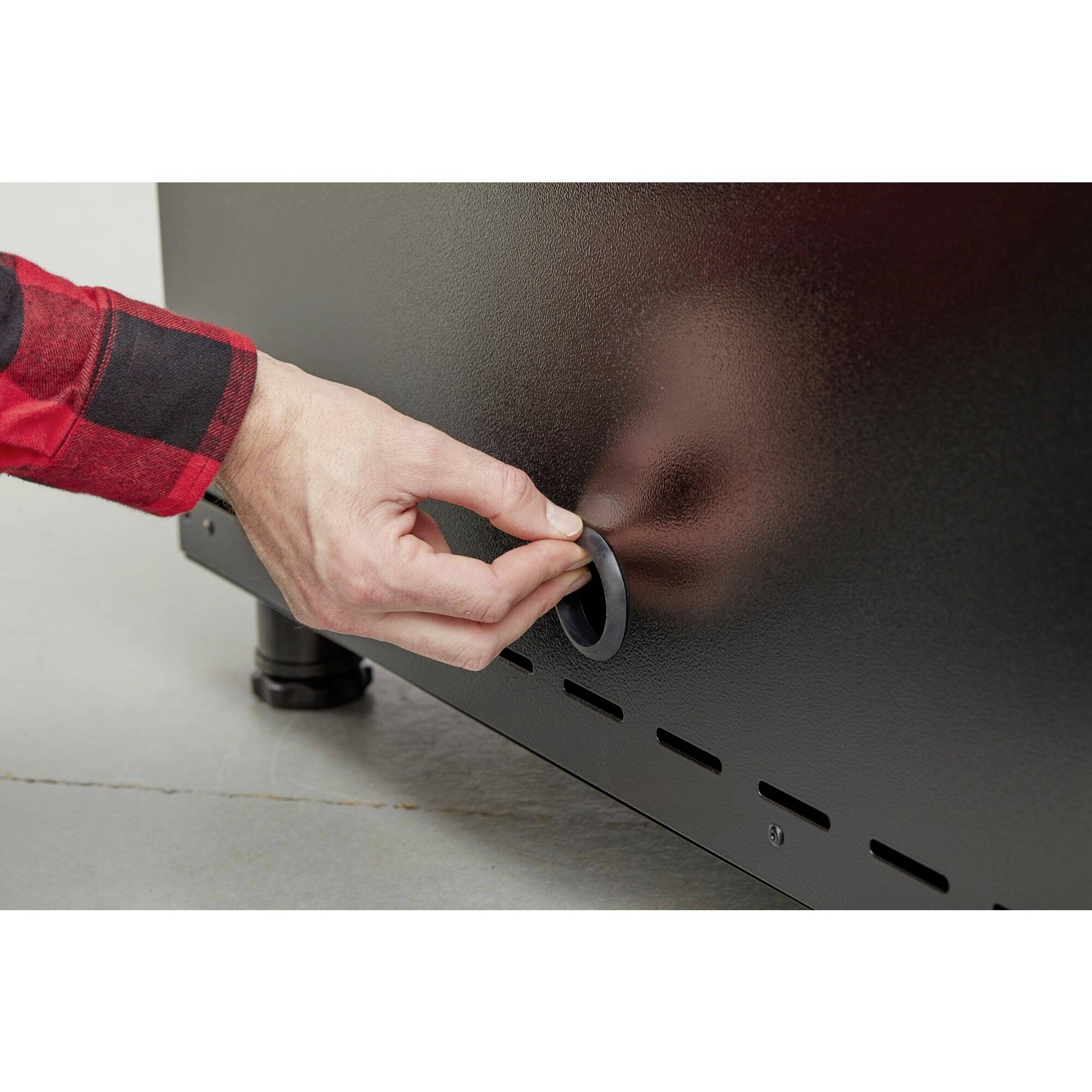 Egy piros ingű kéz egy fekete fémeszköz kerek szellőzőnyílását nyitja egy szürke padlójú helyiségben.