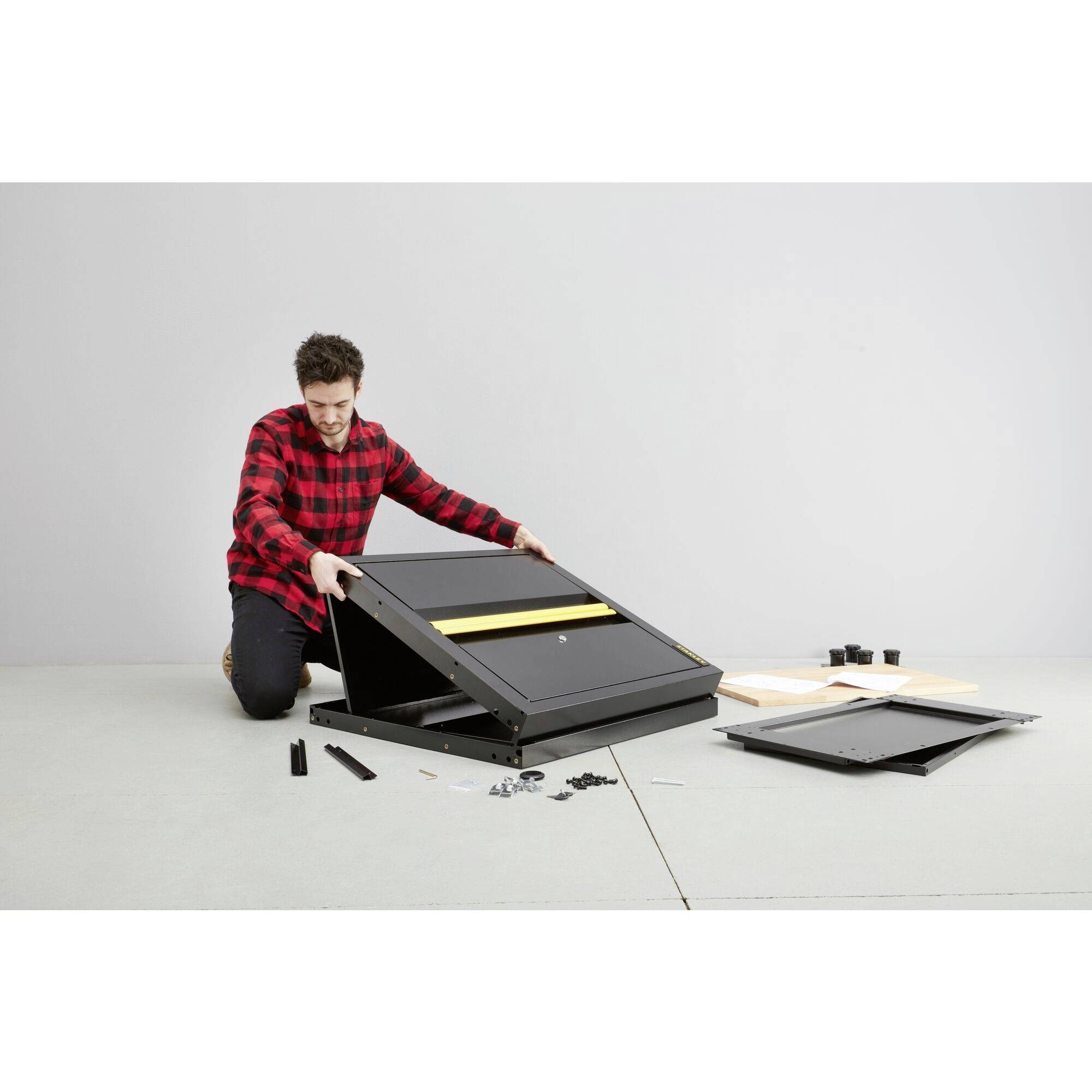 Egy férfi egy fekete bútordarabot szerel össze. Piros-fekete kockás inget visel, és világos padlón térdel. Szerszámok és alkatrészek vannak szétszórva körülötte.