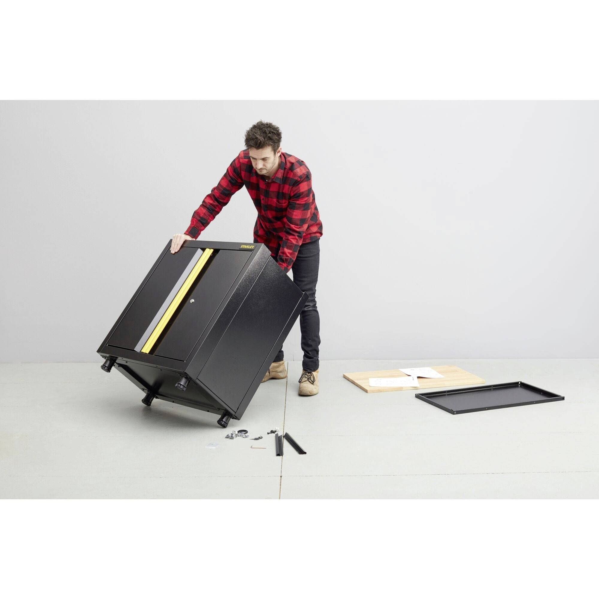 Egy férfi kockás ingben egy fekete fali szekrényt szerel. Szerszámok és útmutatók fekszenek mellette a padlón.