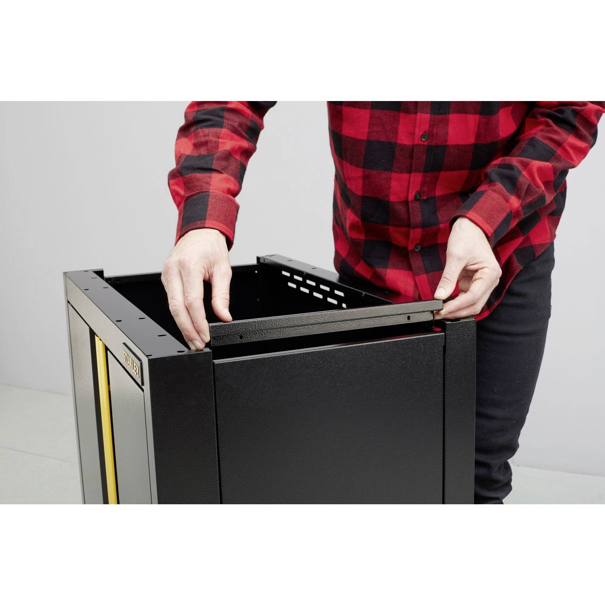 Egy piros-fekete kockás ingű személy egy fekete házat szerel össze, valószínűleg egy műszaki eszközt. A kezek a felső szélét fogják.