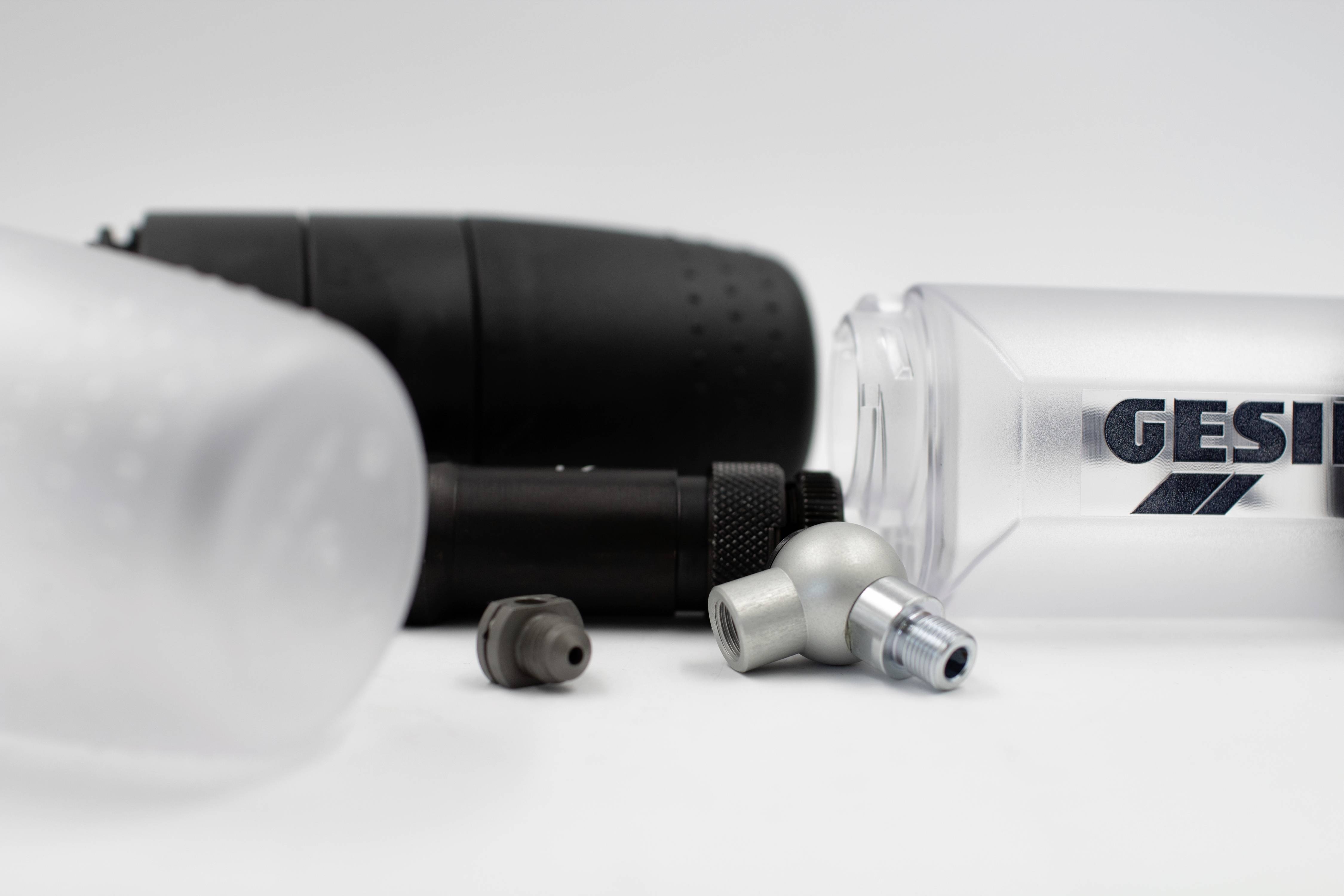 Két ivópalack, egy fekete és egy átlátszó a 'GESIPA' logóval, fekszik egy felületen. Köztük fémdarabok vannak.