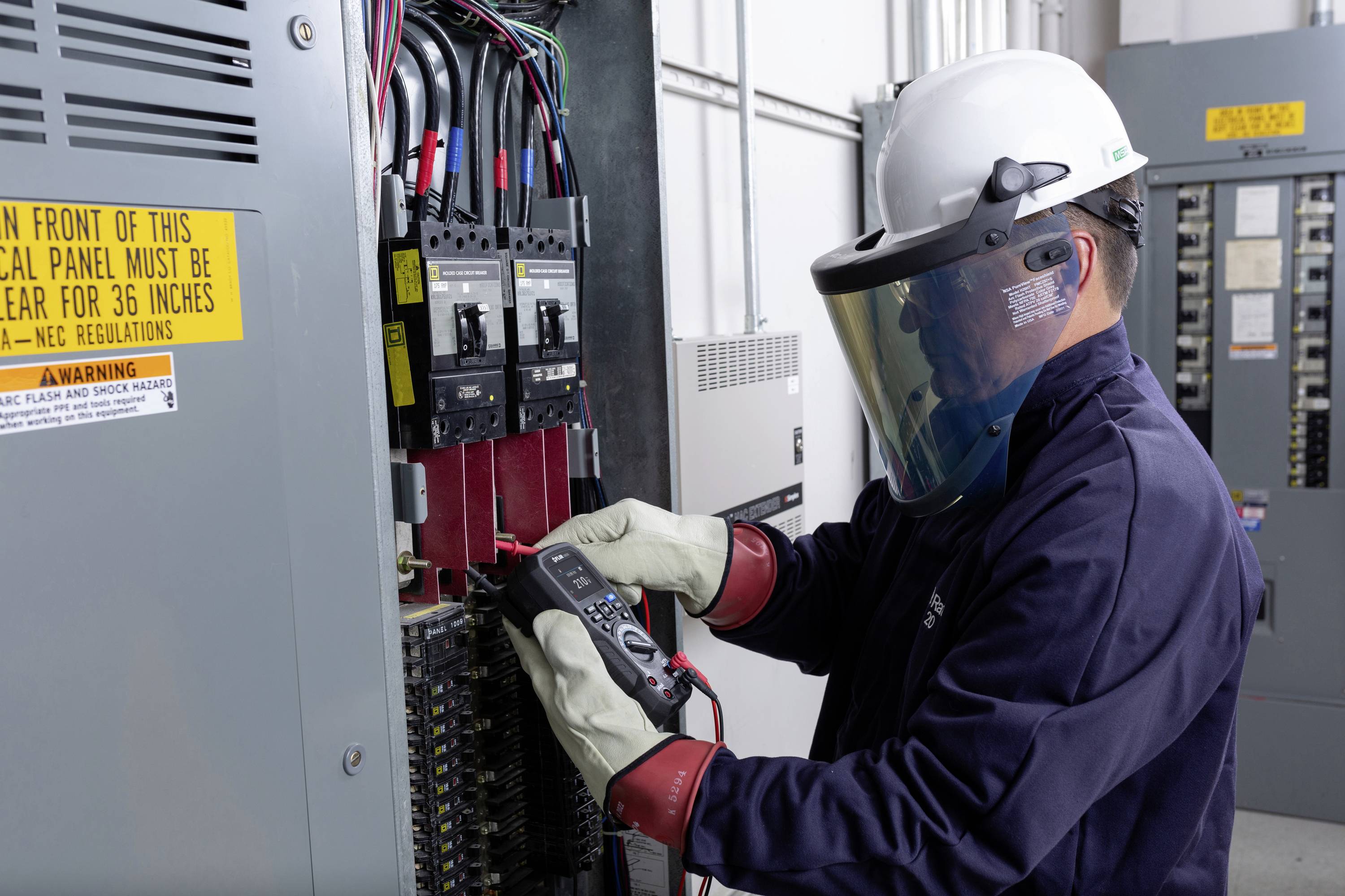 Egy védőruhát viselő technikus egy készülékkel mér egy elektromos elosztószekrényben. Biztonsági figyelmeztetések láthatók.