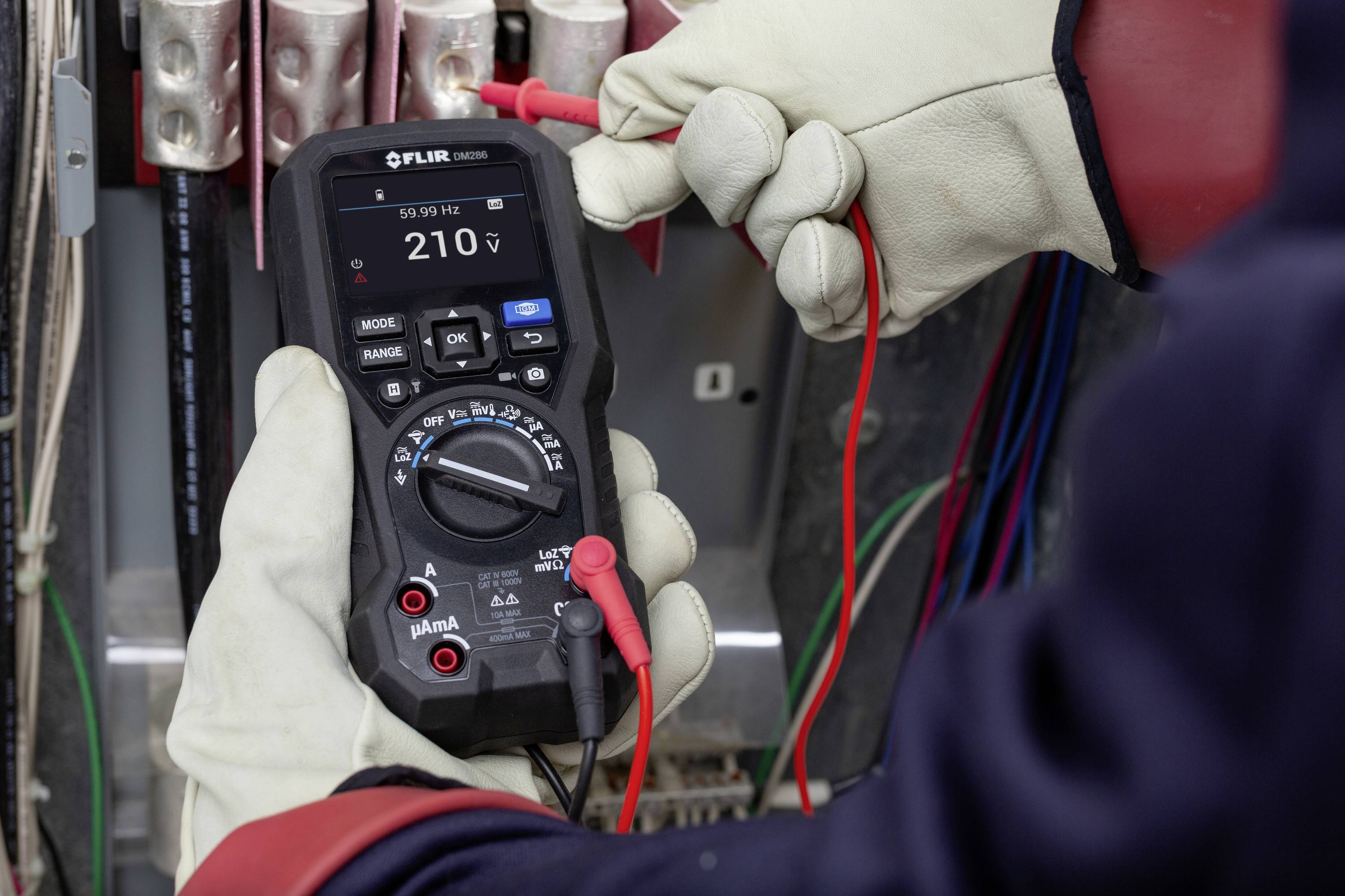 Egy védőkesztyűt viselő személy digitális multiméterrel méri a feszültséget egy elektromos elosztószekrényben.