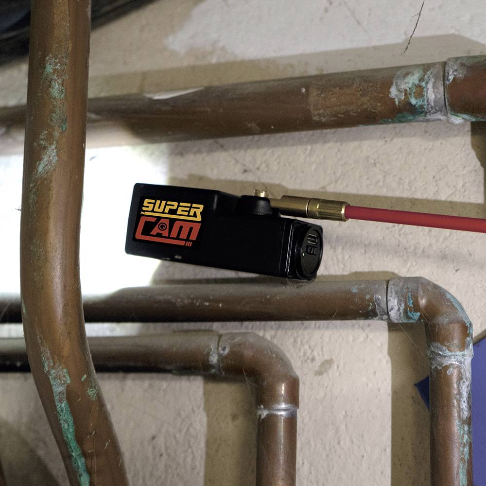 Egy 'Super Cam' feliratú kamera egy piros rúdra erősítve, csövek által körülvéve egy ipari térben.