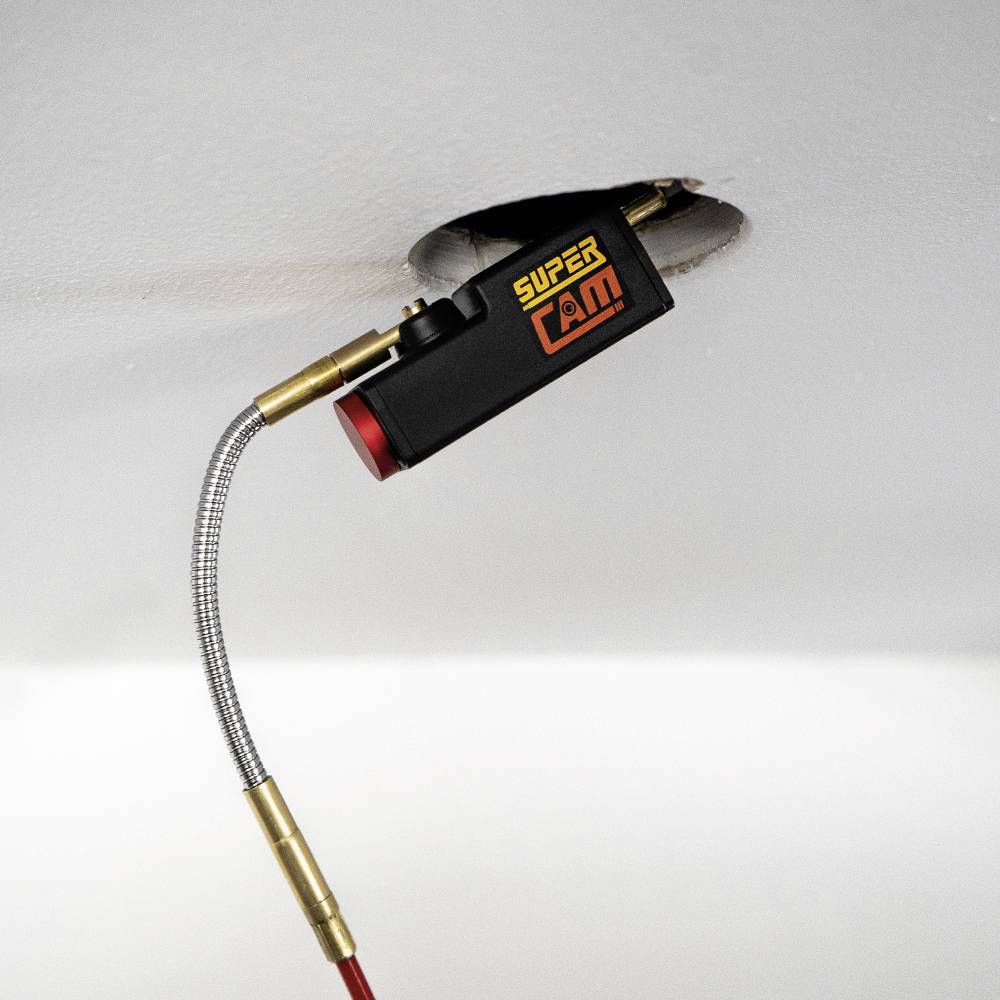 Egy fekete kamera, amelyen 'SUPERCAM' felirat látható, egy fehér mennyezeten lévő lyukon át lóg be, egy rugalmas kábellel rögzítve.