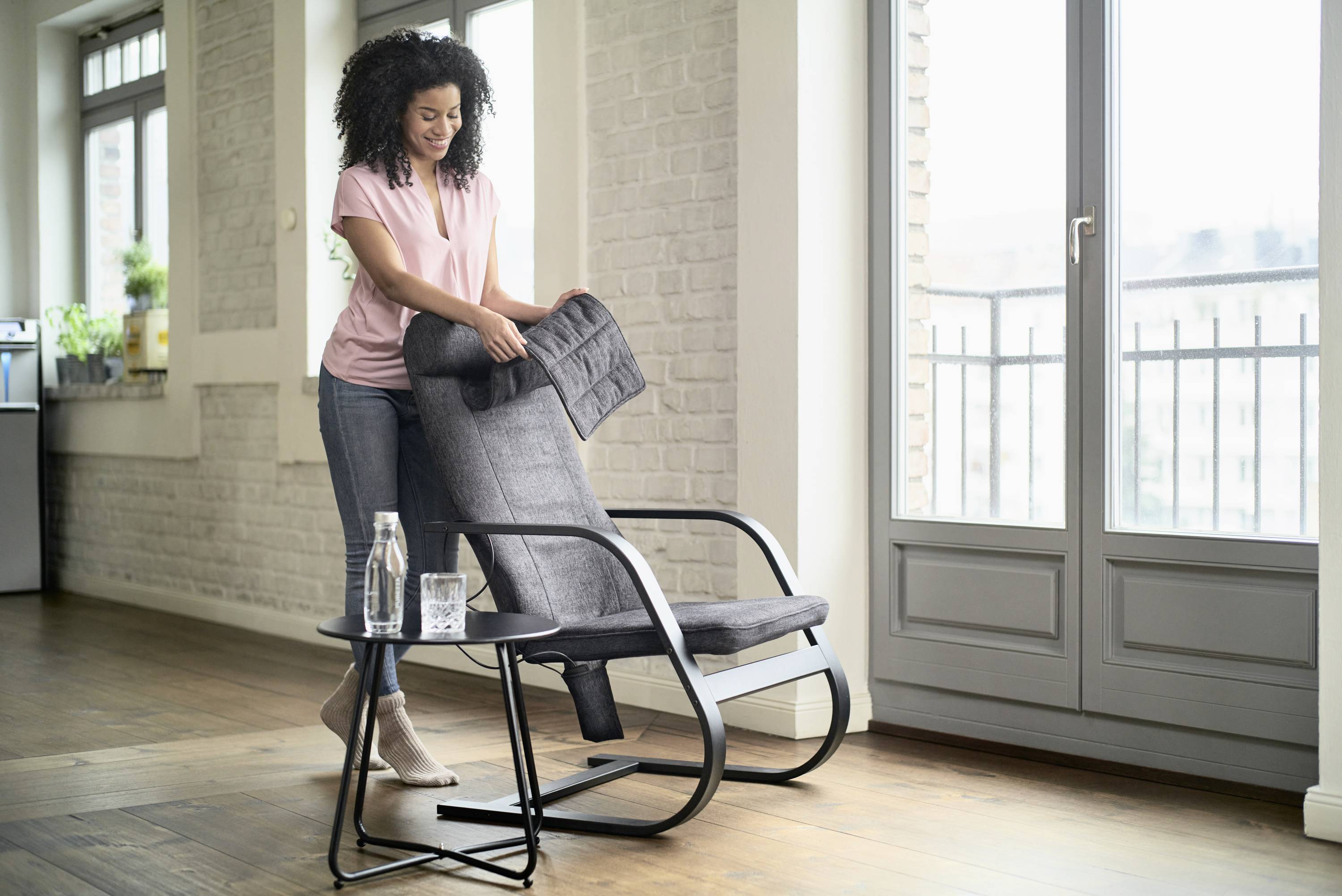 Egy nő elrendez egy ülőpárnát egy modern hintaszéken egy világos nappaliban, nagy ablakok előtt. Mellette egy asztal áll egy pohár vízzel.