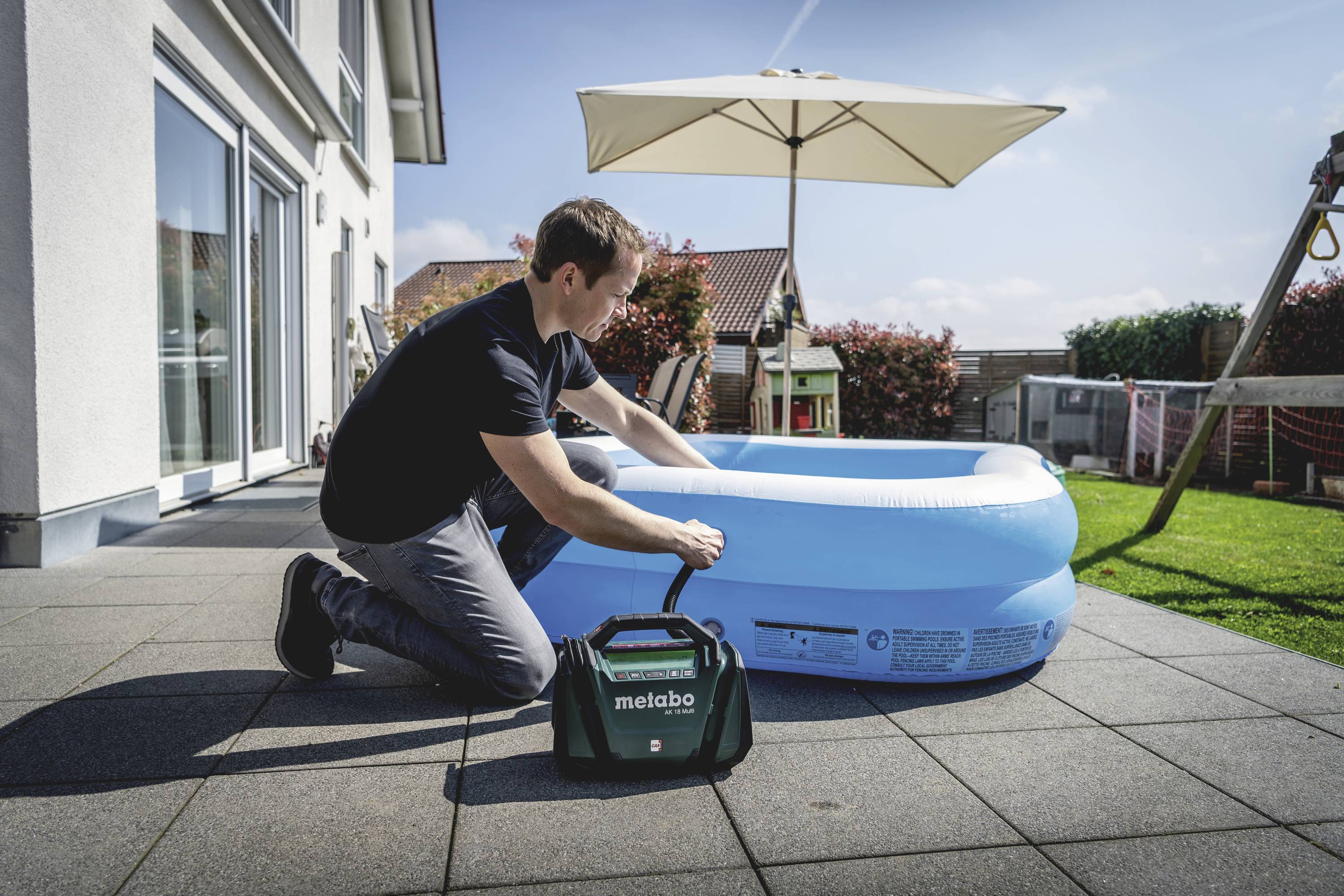 Egy férfi egy elektromos pumpával felfújja a kerek, felfújható gyerekmedencét egy teraszon. A háttérben egy napnapernyős kert látható.