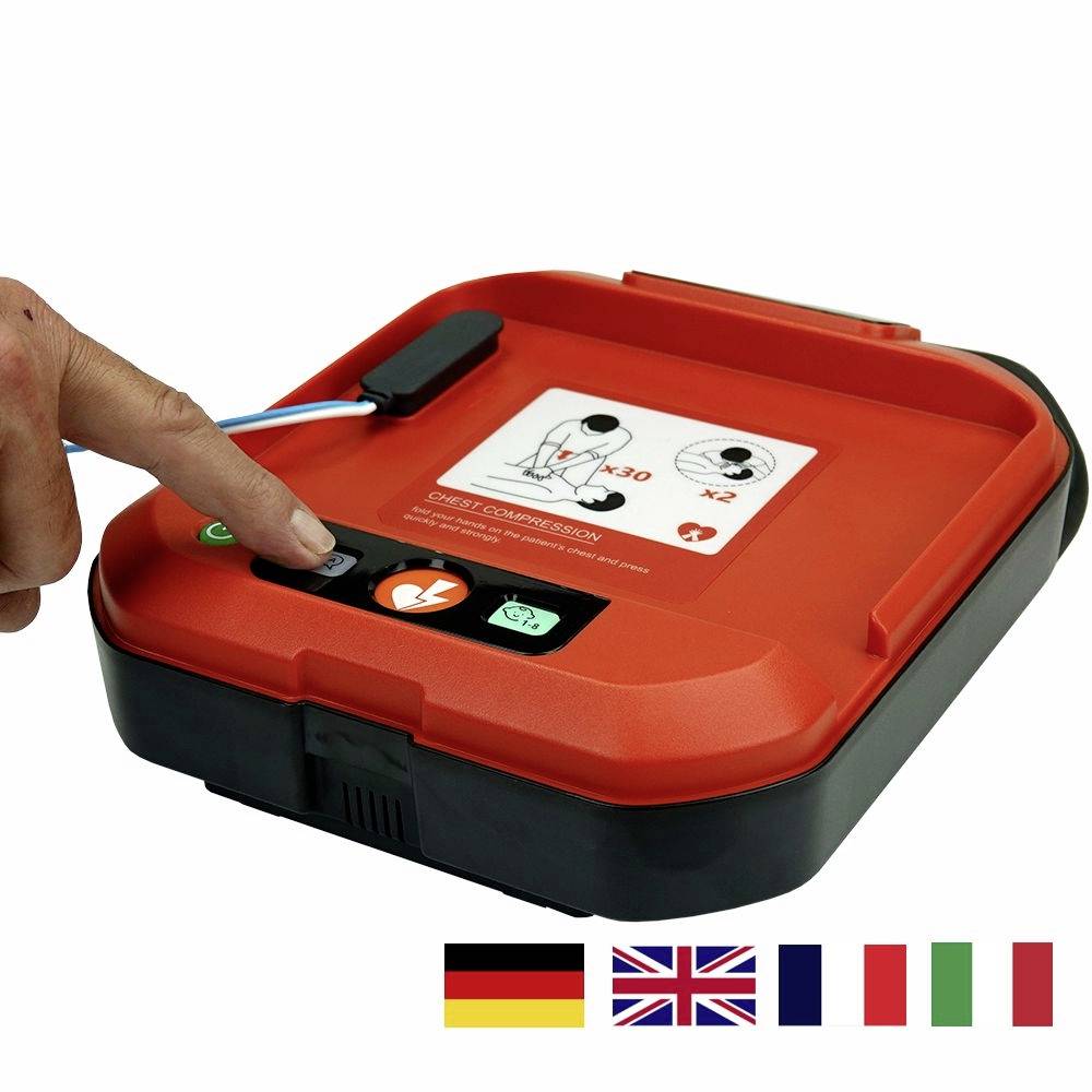 Egy piros defibrillátor elsősegélynyújtáshoz, amelyen német, brit, francia és olasz zászlók láthatók. Egy kéz nyomja a bekapcsológombot.
