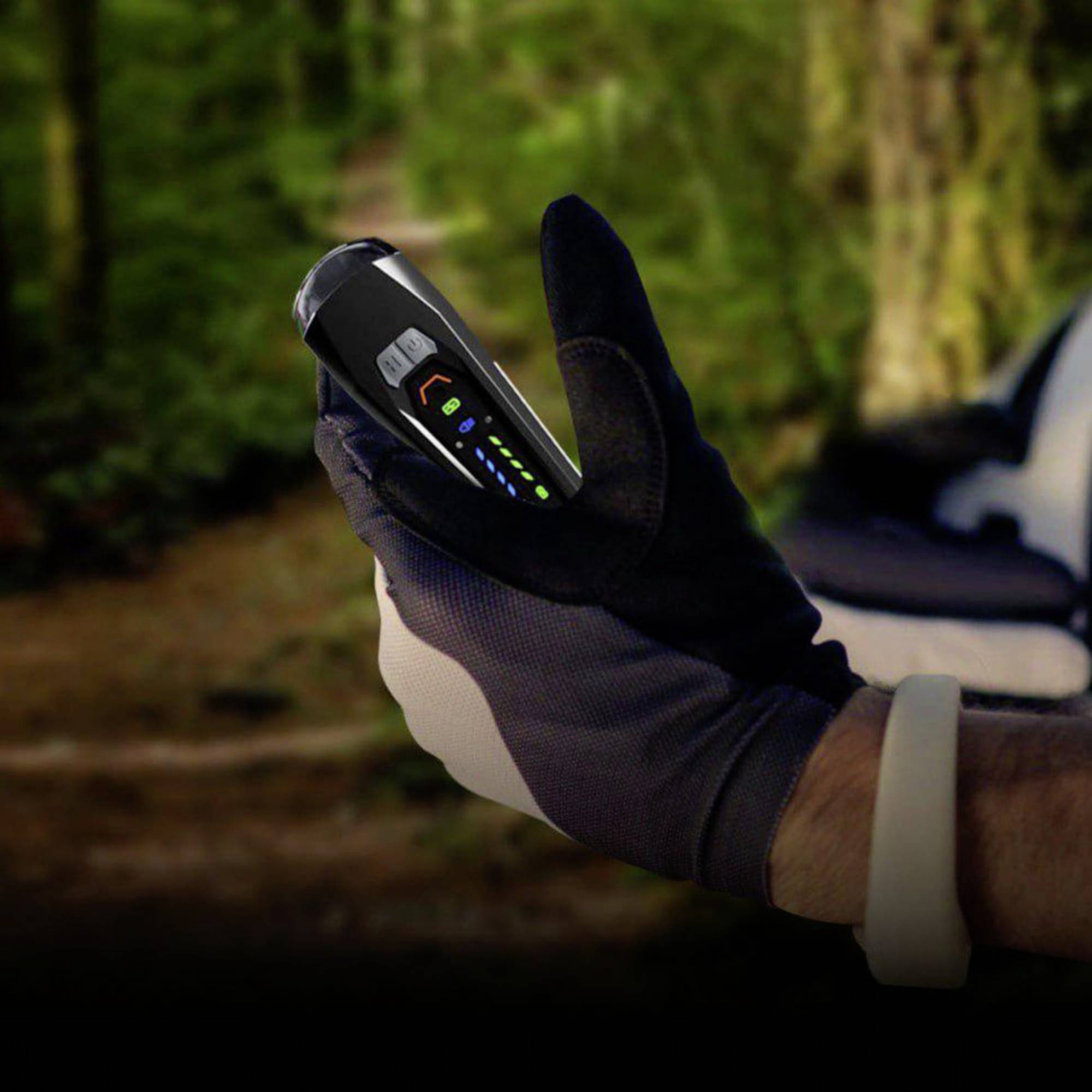 Egy védőkesztyűs kéz tart egy hordozható készüléket több világító kijelzővel. A háttér elmosódott, valószínűleg erdei ösvény.