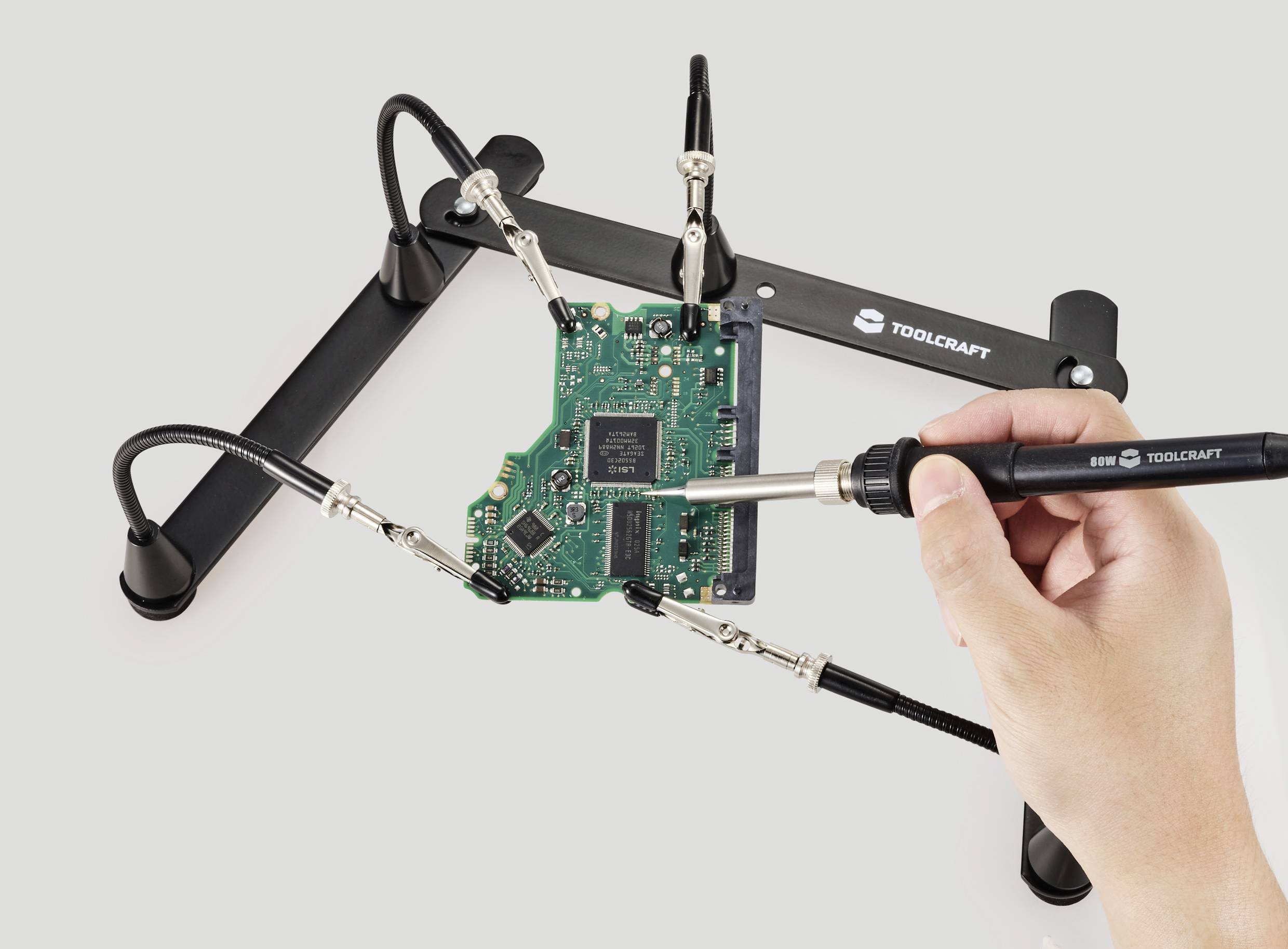 Egy kéz egy zöld nyomtatott áramkört forraszt, amelyet egy tartóban rögzítettek. A háttérben egy TOOLCRAFT logó látható.