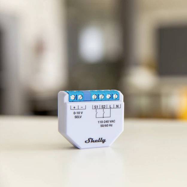 Egy kisméretű, fehér elektronikai eszköz csatlakozókkal, amelyen a '0-10 V SELV 50/60 Hz Shelly' felirat olvasható, homályos háttérrel.