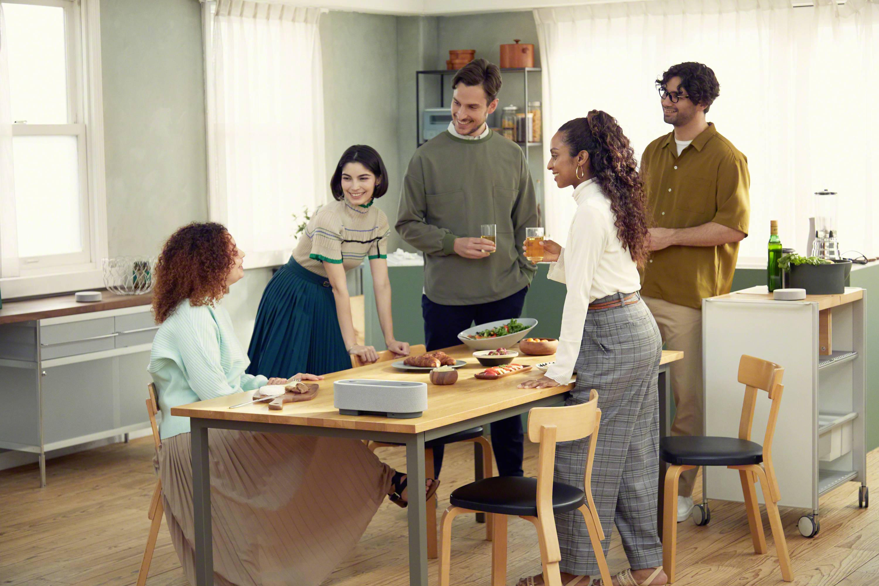 Öt ember áll egy világos helyiségben egy asztal körül, amelyen étel és italok vannak. Úgy tűnik, nevetgélnek és oldottan beszélgetnek.