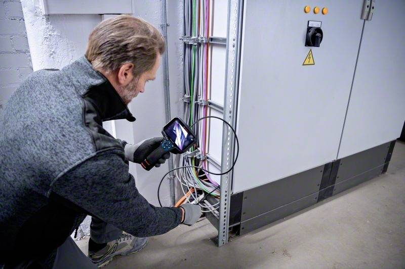 Egy férfi egy kameraeszköz segítségével ellenőrzi az elektromos kábeleket egy elosztószekrényben, hogy részletes képet kapjon a kábel csatlakozásokról.