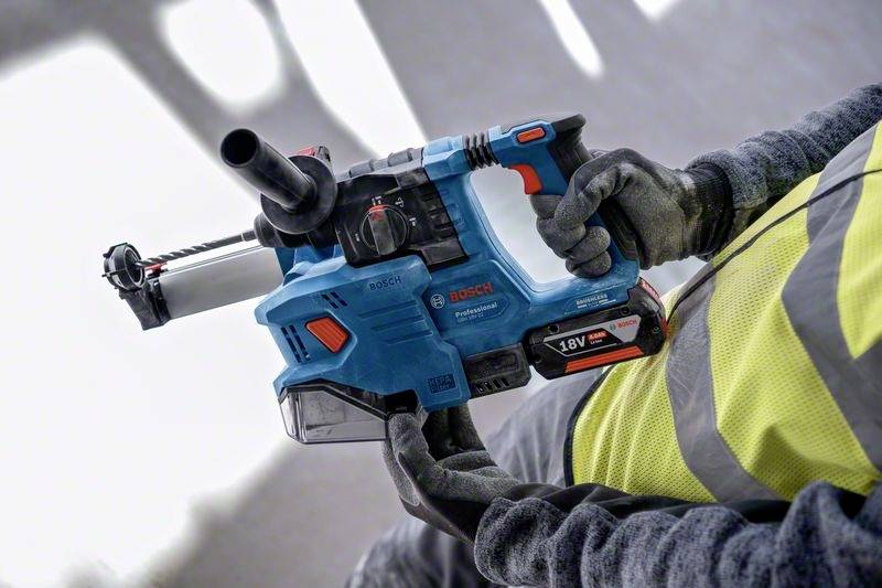Egy munkásruhát viselő személy egy kék Bosch elektromos fúrógépet tart 18V-os akkumulátorral, használatra készen.