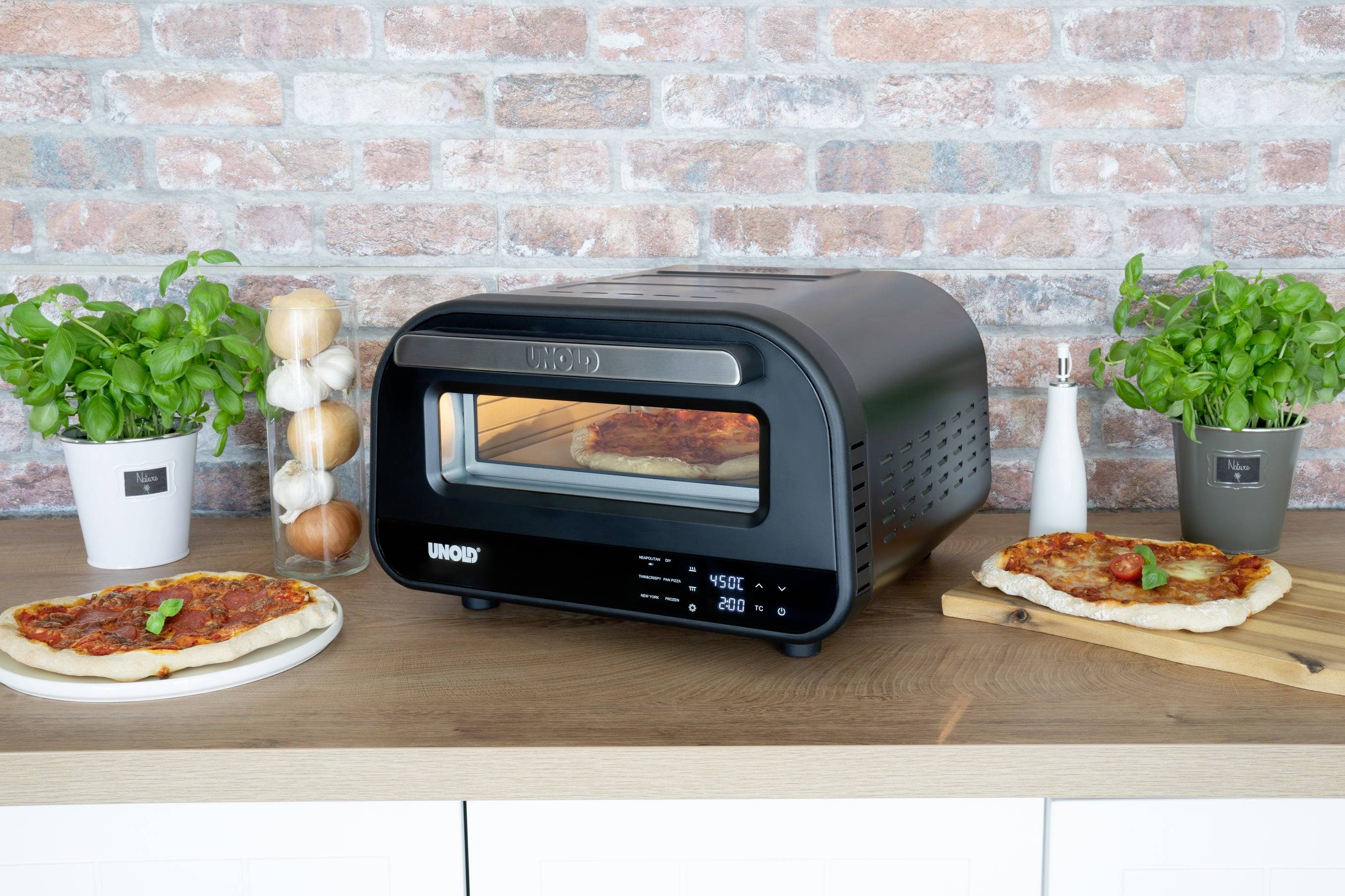 Egy fekete pizzasütő egy fadeszkán áll, körülvéve két frissen sült pizzával, bazsalikom növényekkel és fűszerekkel.