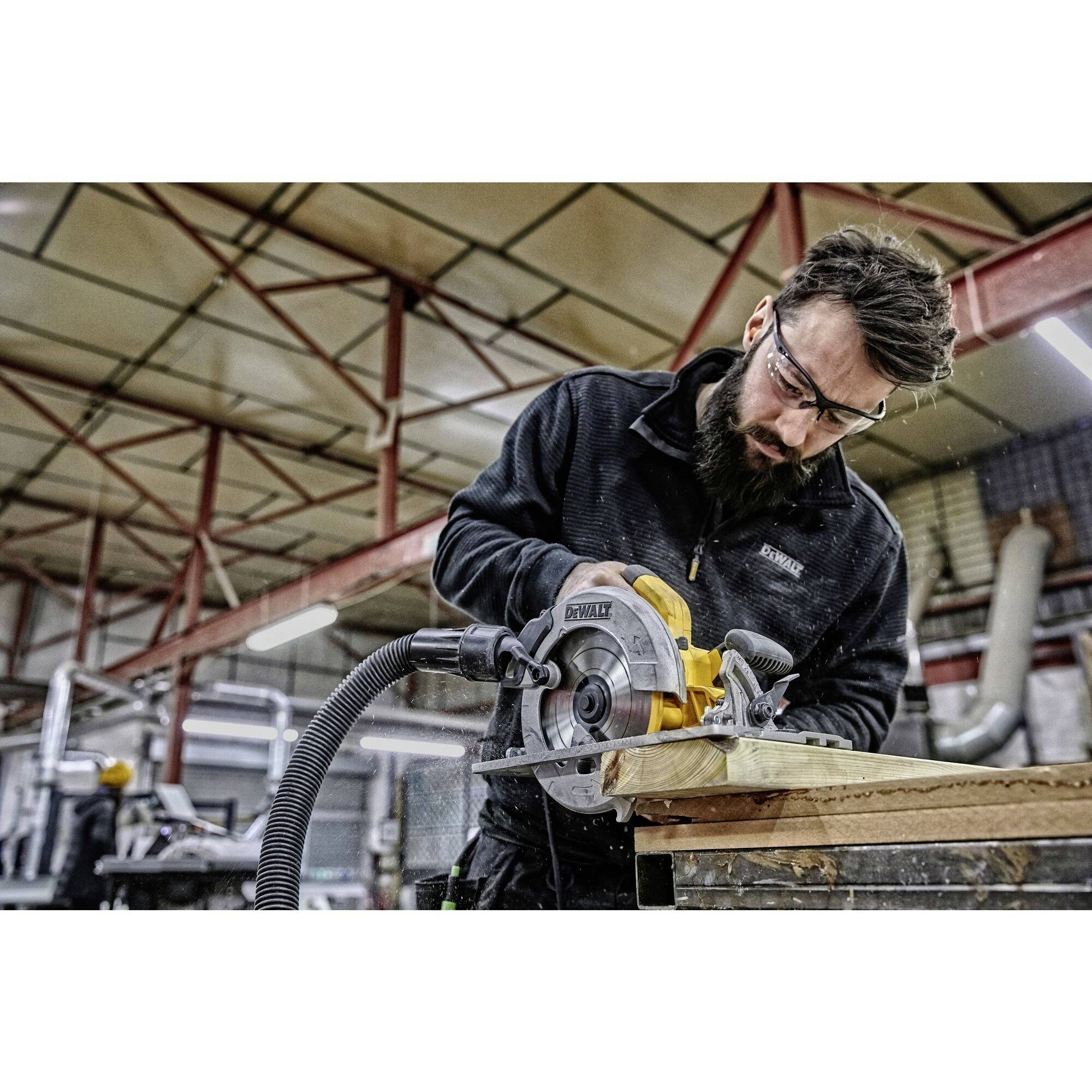 Egy férfi védőszemüvegben elektromos fűrésszel dolgozik egy műhelyben. Egy falapot vág egy munkapadon.