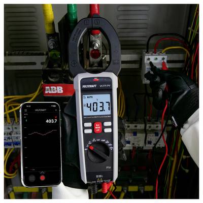 A technikus egy csipesz ampermérővel és egy okostelefonnals mér elektromos feszültséget egy panelen, mindkettő körülbelül 403,7 V értéket mutat.
