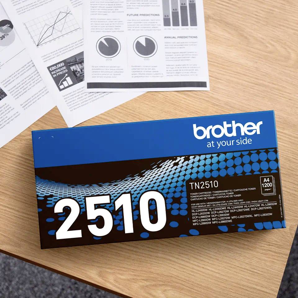 Egy fa íróasztalon egy 'Brother TN2510' tonerkazetta fekszik szétterített dokumentumok, grafikák és szövegek mellett.